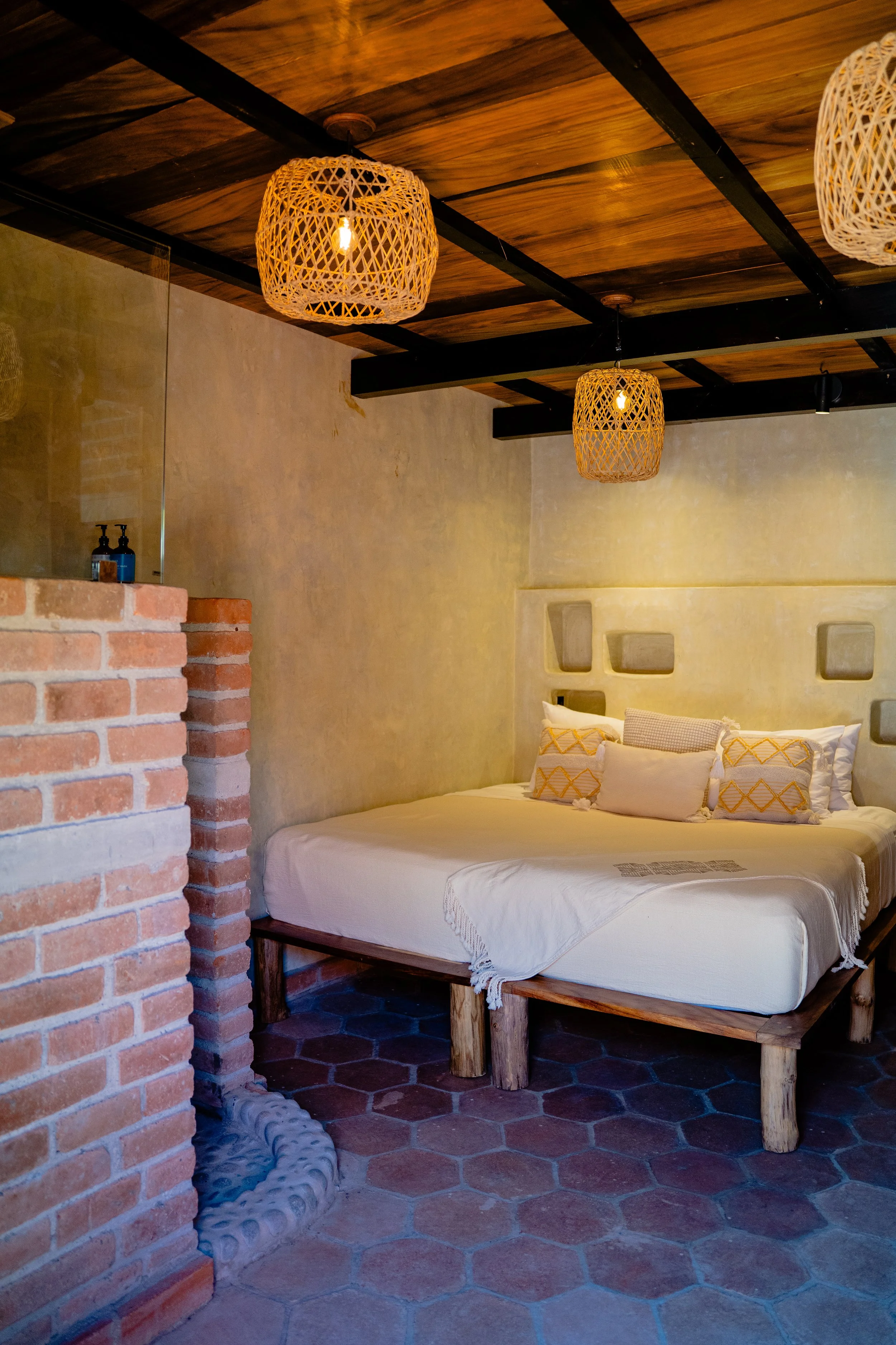 A bedroom with a rustic style, featuring a low bed with white bedding and decorative pillows, a wooden headboard with built-in shelves, a brick wall in the corner, a textured concrete wall painted in neutral tones, hanging woven wicker pendant lights, and a brick and tile floor.