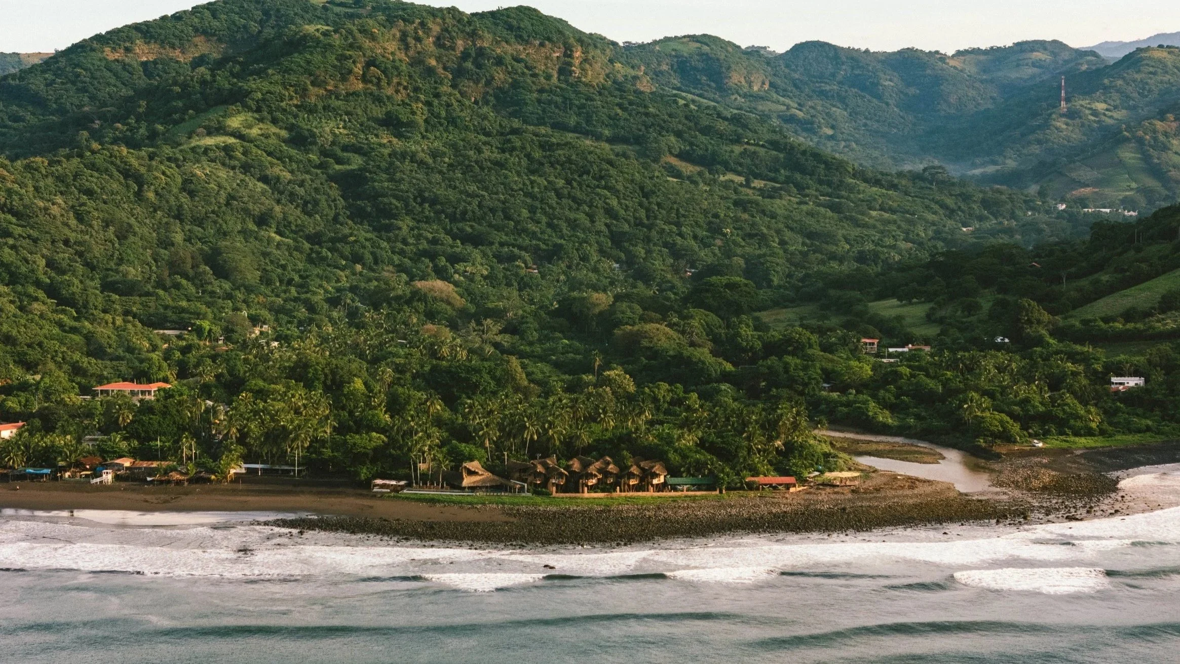Beachfront with a small river flowing into the ocean, dense green forested hills in the background, and a few houses and huts along the shoreline.