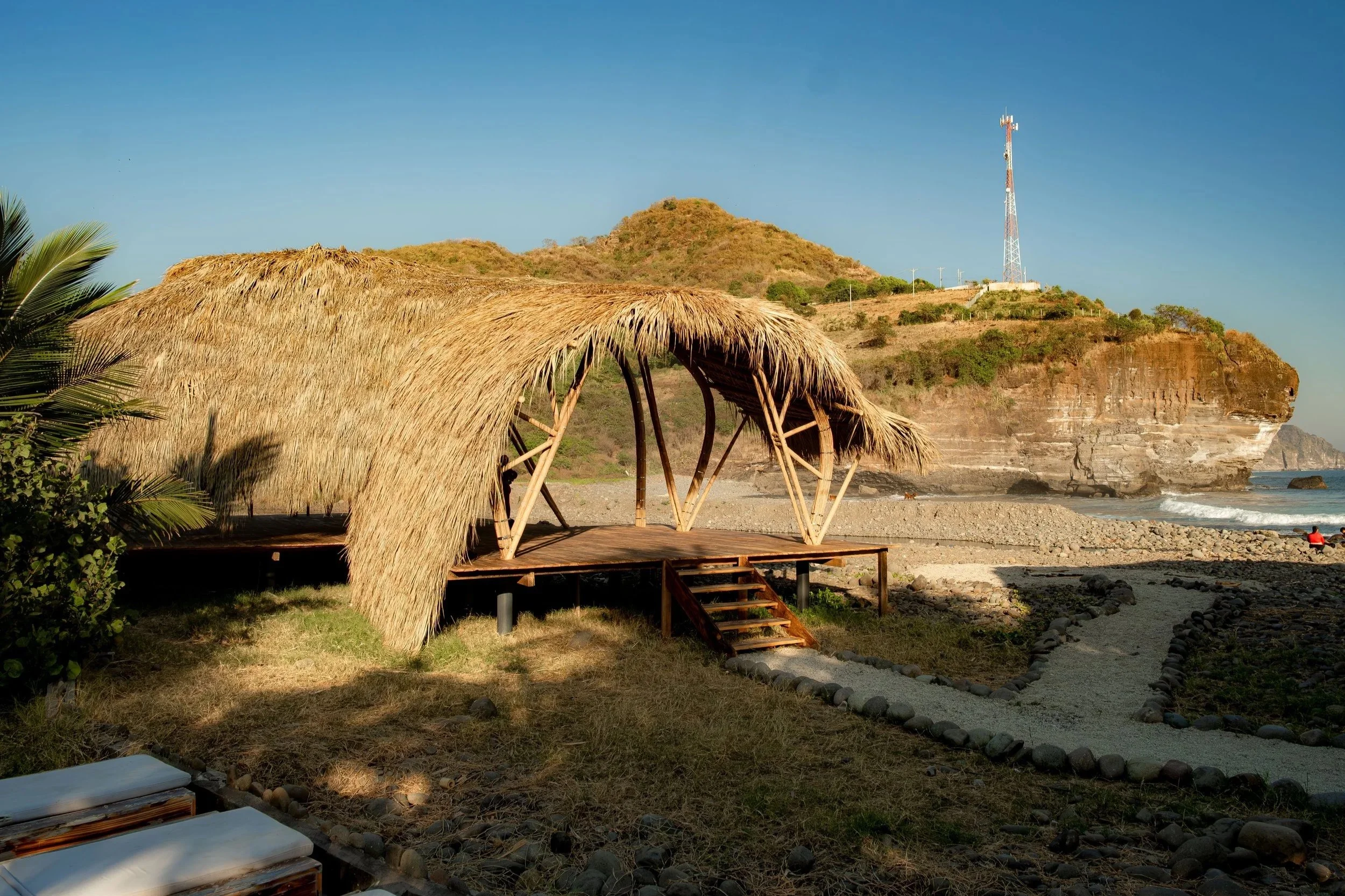 A beach scene with a thatched-roof hut, rocky shoreline, and a cliff in the background with a communications tower on top. There is one person sitting near the water and a person walking along the beach.