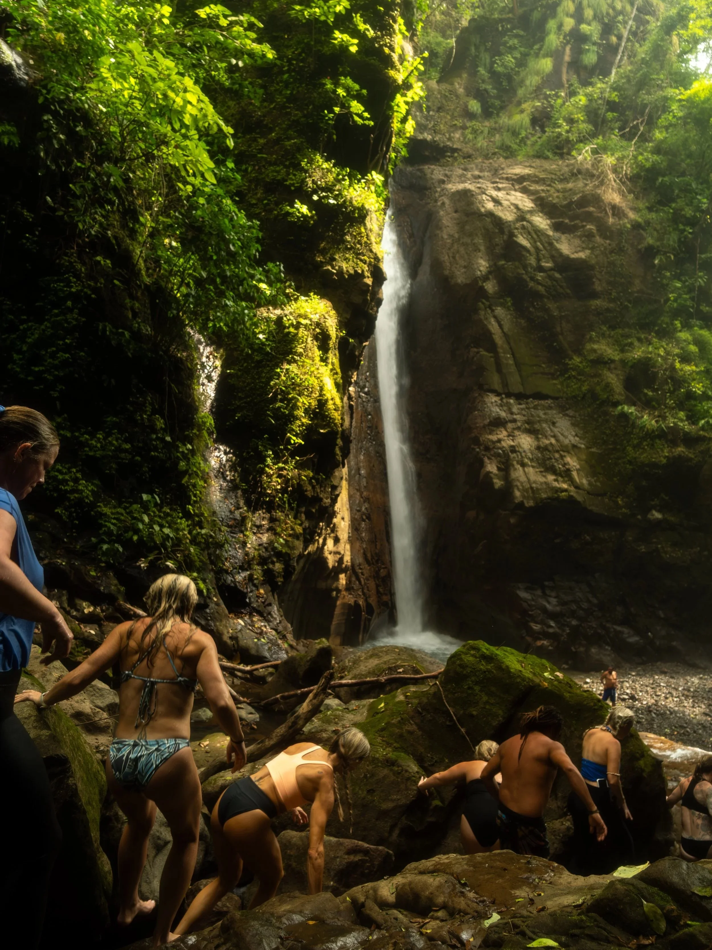 People, mostly women, in swimsuits, standing on rocks and bending over in a stream, with a lush green forest and a tall waterfall in the background.