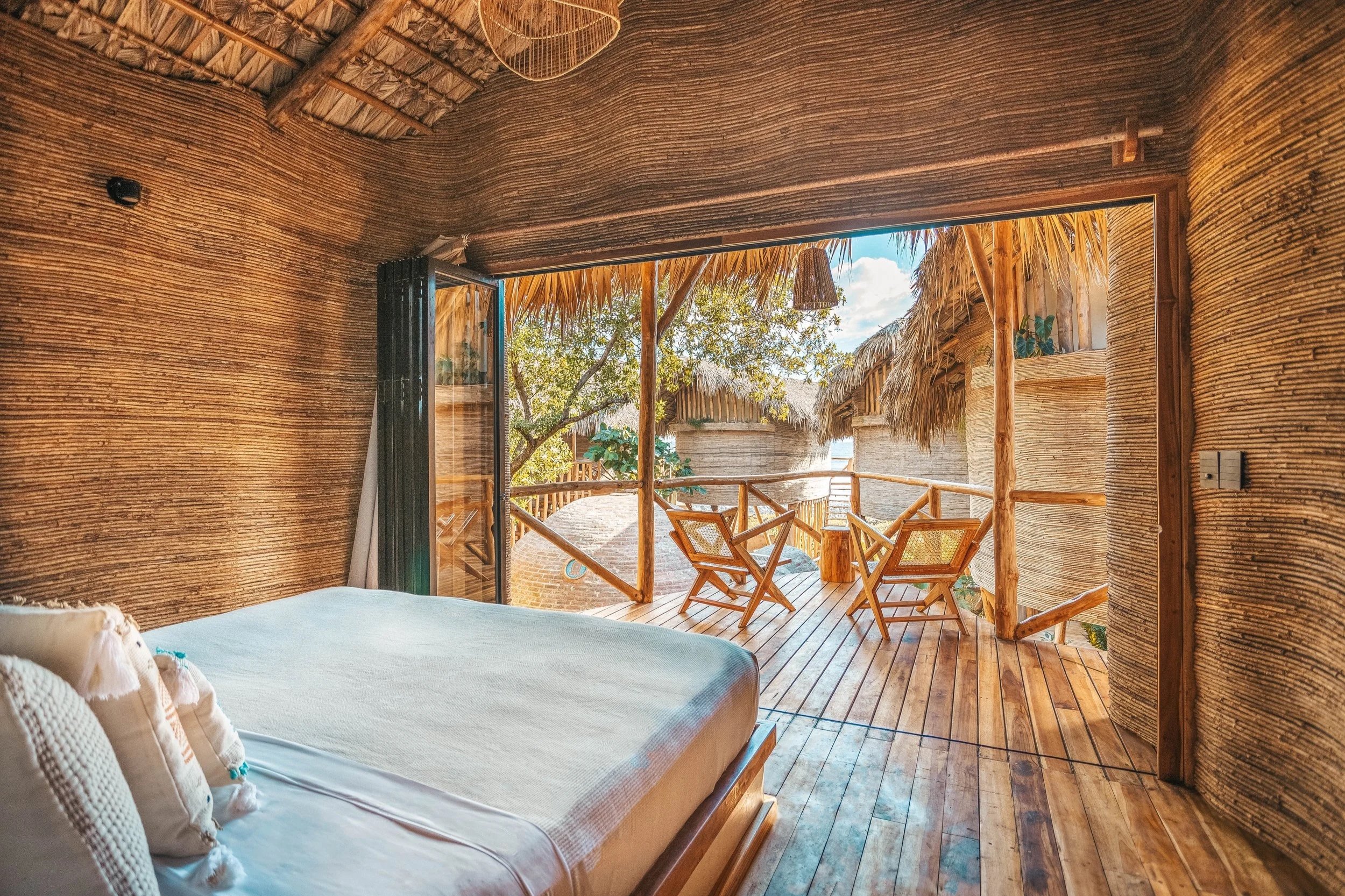 Room with wooden walls, open sliding door leading to a balcony with wooden chairs, and thatched roof outside. Seen daylight.