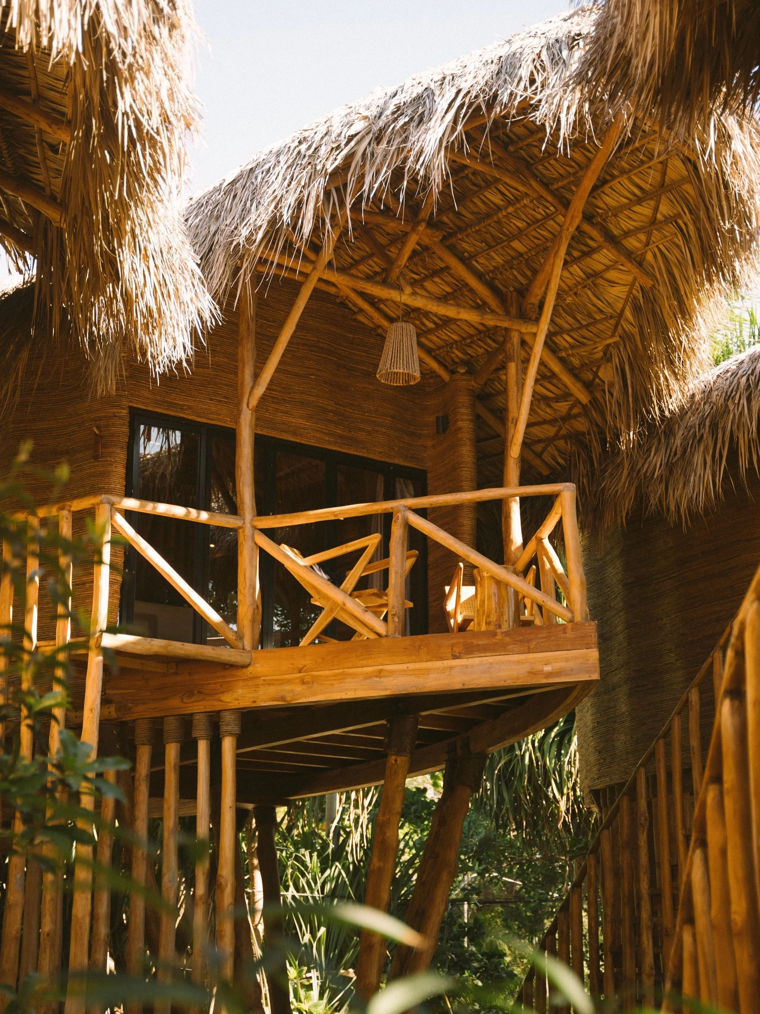 A tropical hut with a thatched roof made of straw, supported by wooden poles, with a balcony and hanging lamp, surrounded by lush greenery.