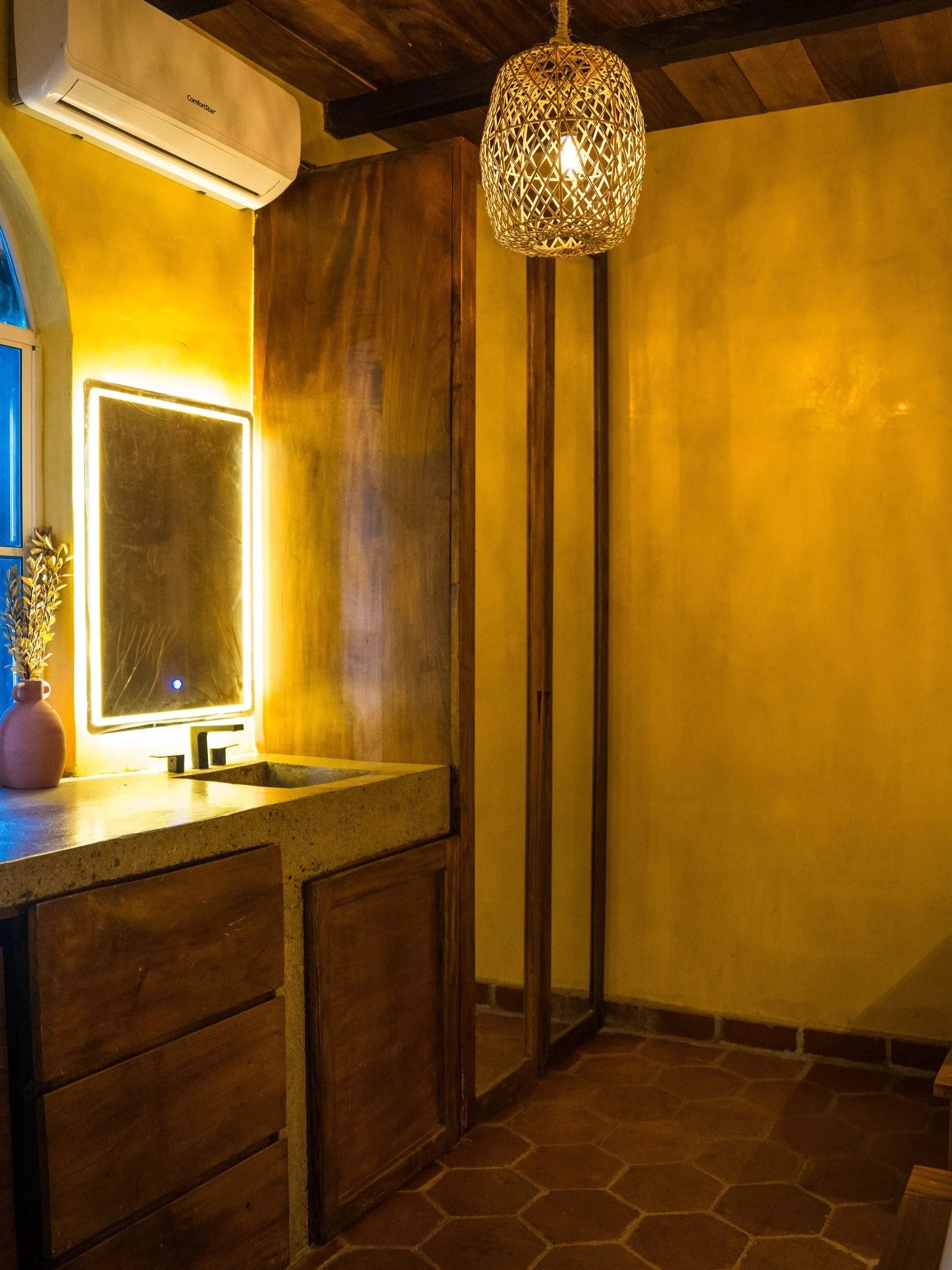 Interior of a room with yellow painted walls, a wooden cabinet, a mirror with backlighting, a potted plant with dried flowers, a wicker hanging light fixture, and an air conditioning unit on the wall.