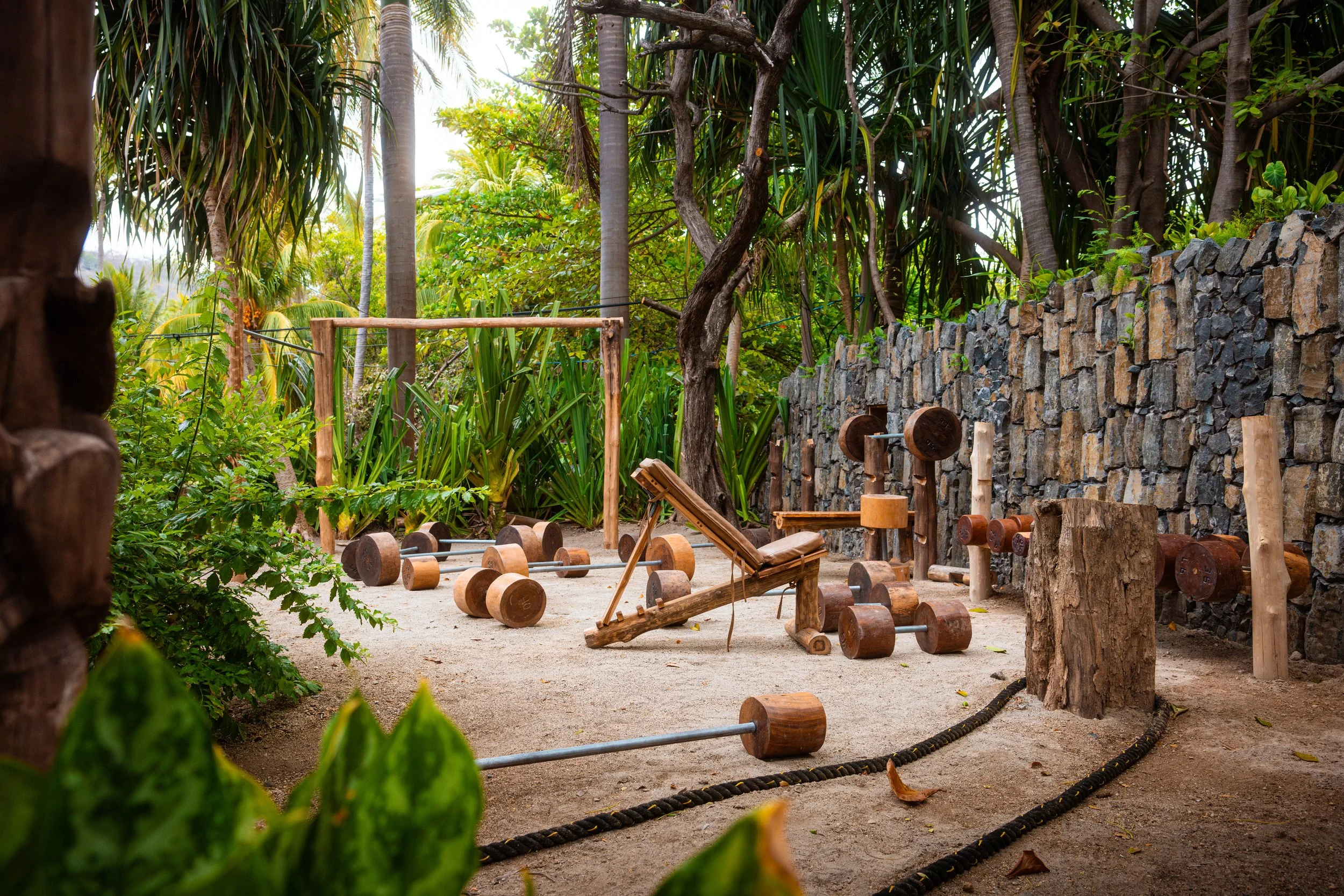 Outdoor fitness area with scattered wooden weights, benches, and ropes amid lush green plants and trees surrounded by a stone wall.