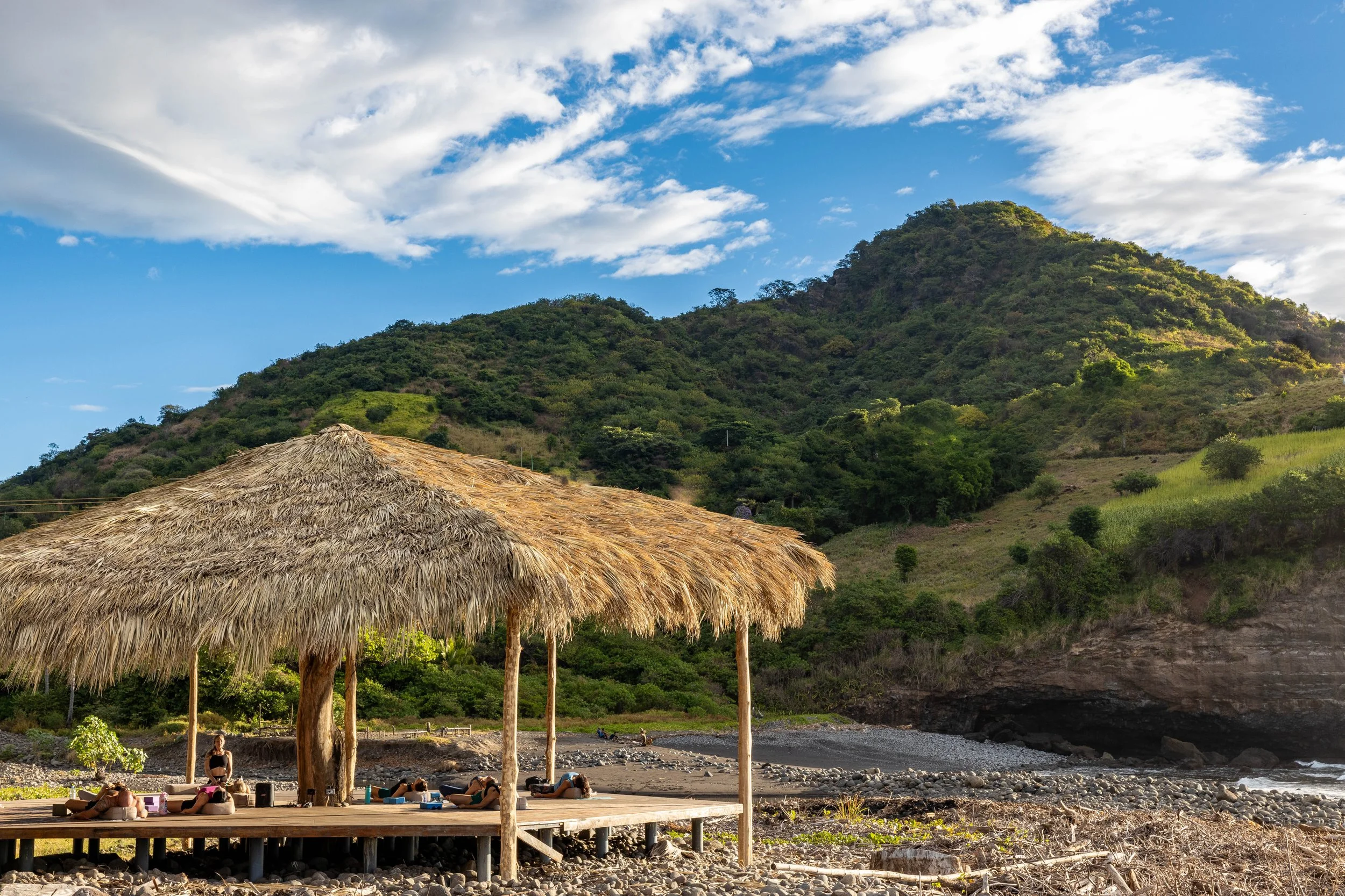 A group of people lying on mats under a thatched roof gazebo on a rocky beach with lush green hills and a partly cloudy sky in the background.