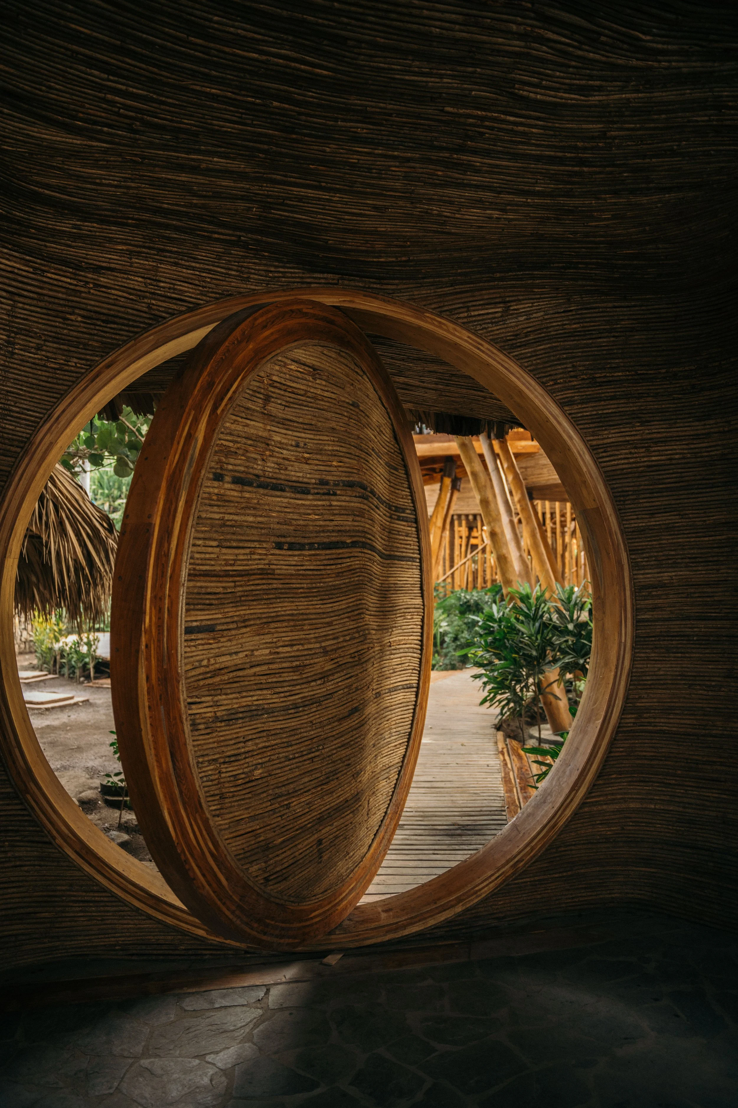 Close-up of a round mirror with a wooden frame, mounted on a wall with dark textured surface, reflecting a pathway with plants, wooden buildings, and greenery outside.