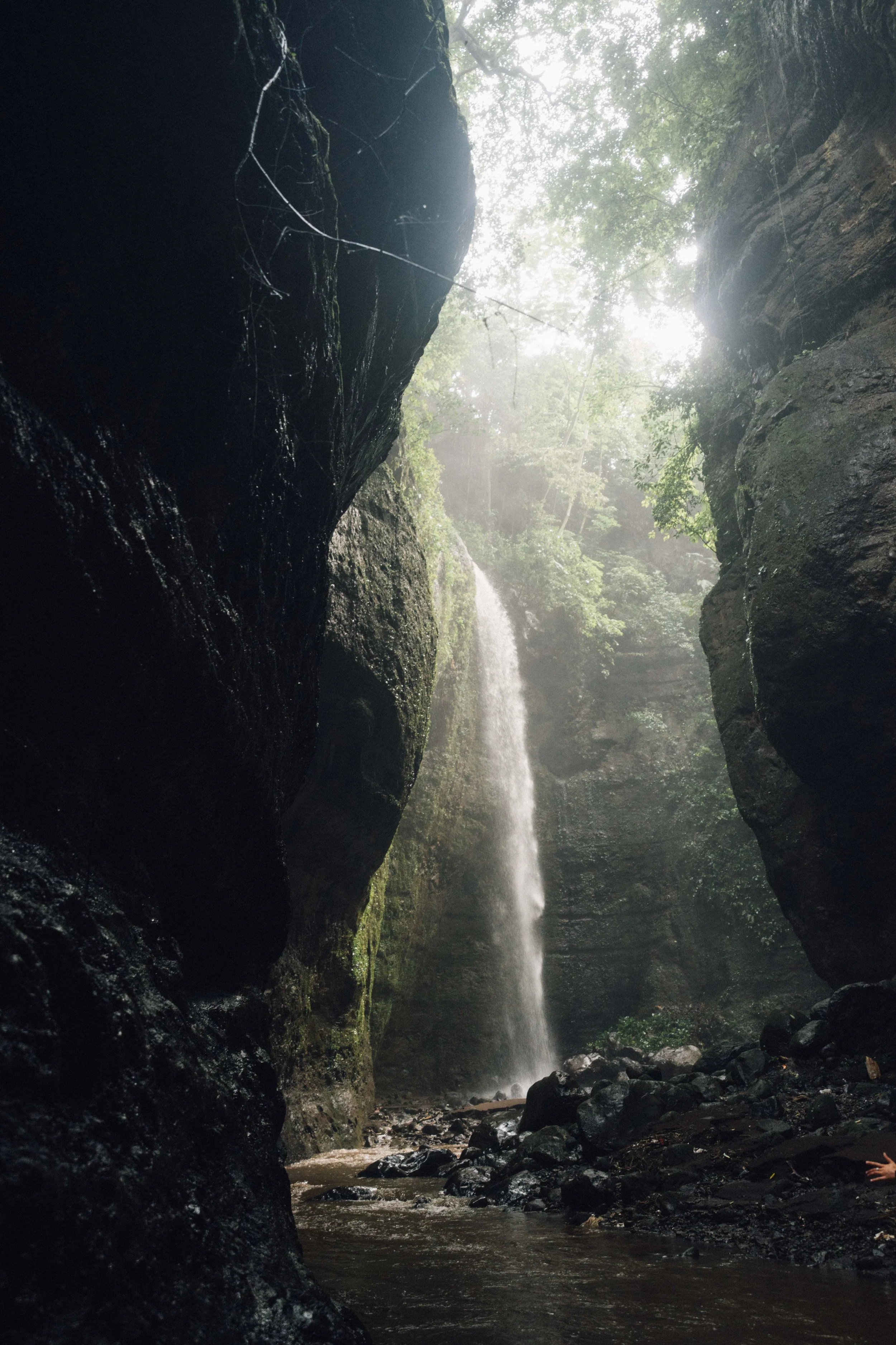 A view of a waterfall in a canyon with steep rock walls, greenery, and mist or spray from the waterfall, with water flowing at the bottom over rocks.