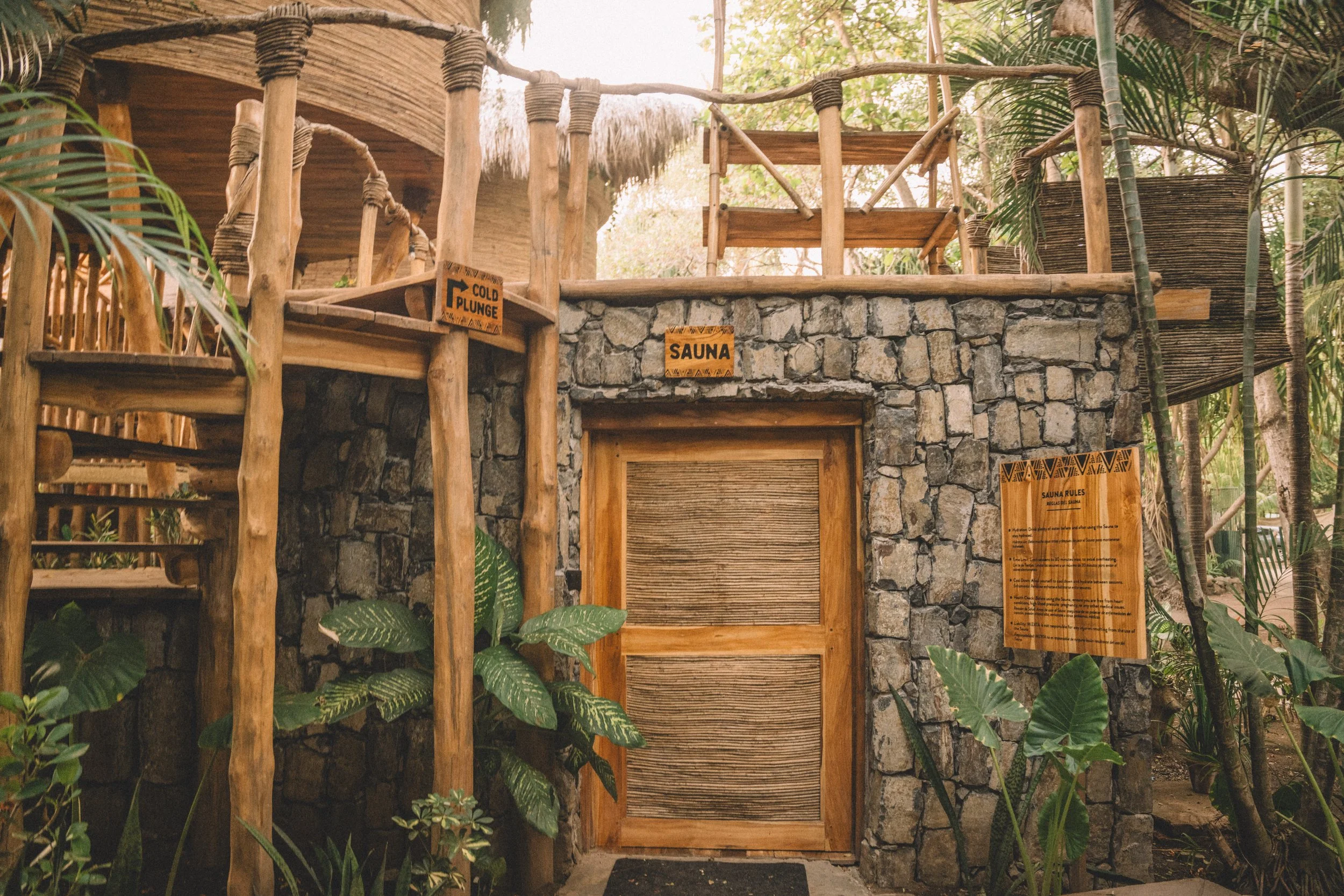 Wooden sauna entrance next to a stone wall with signs indicating sauna rules and regulations, surrounded by tropical plants.
