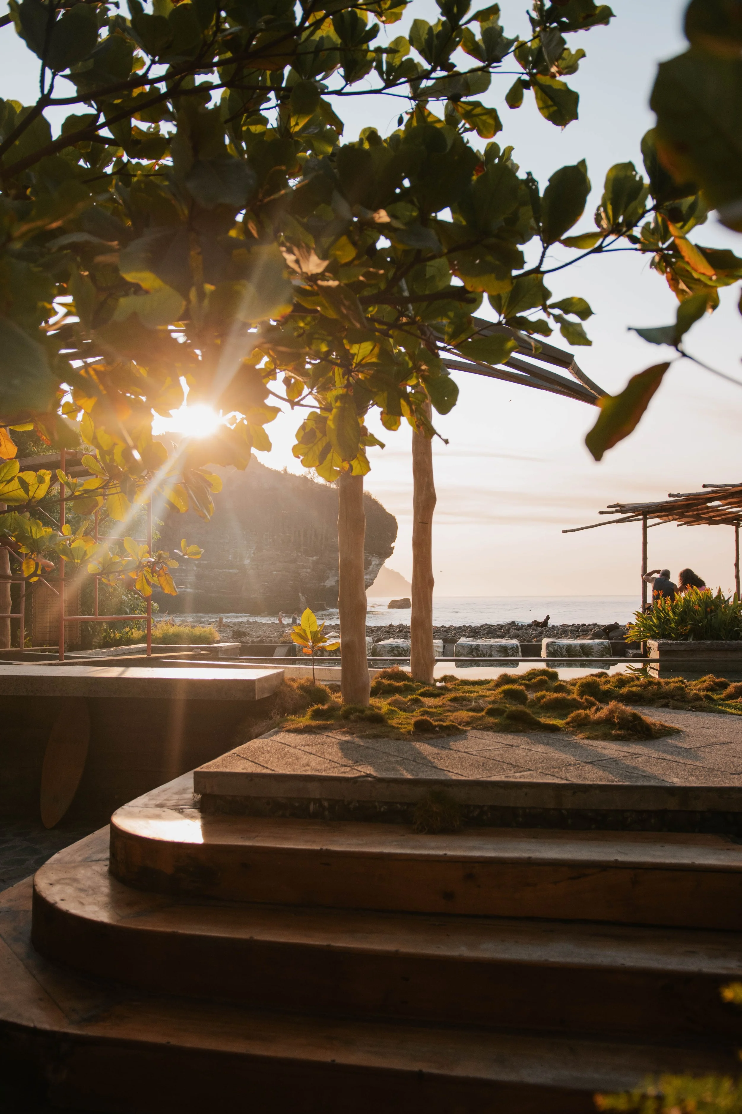 Sunset view of a beach with trees, rocky shoreline, and a small thatched hut or structure, with a person sitting near the edge of the water.