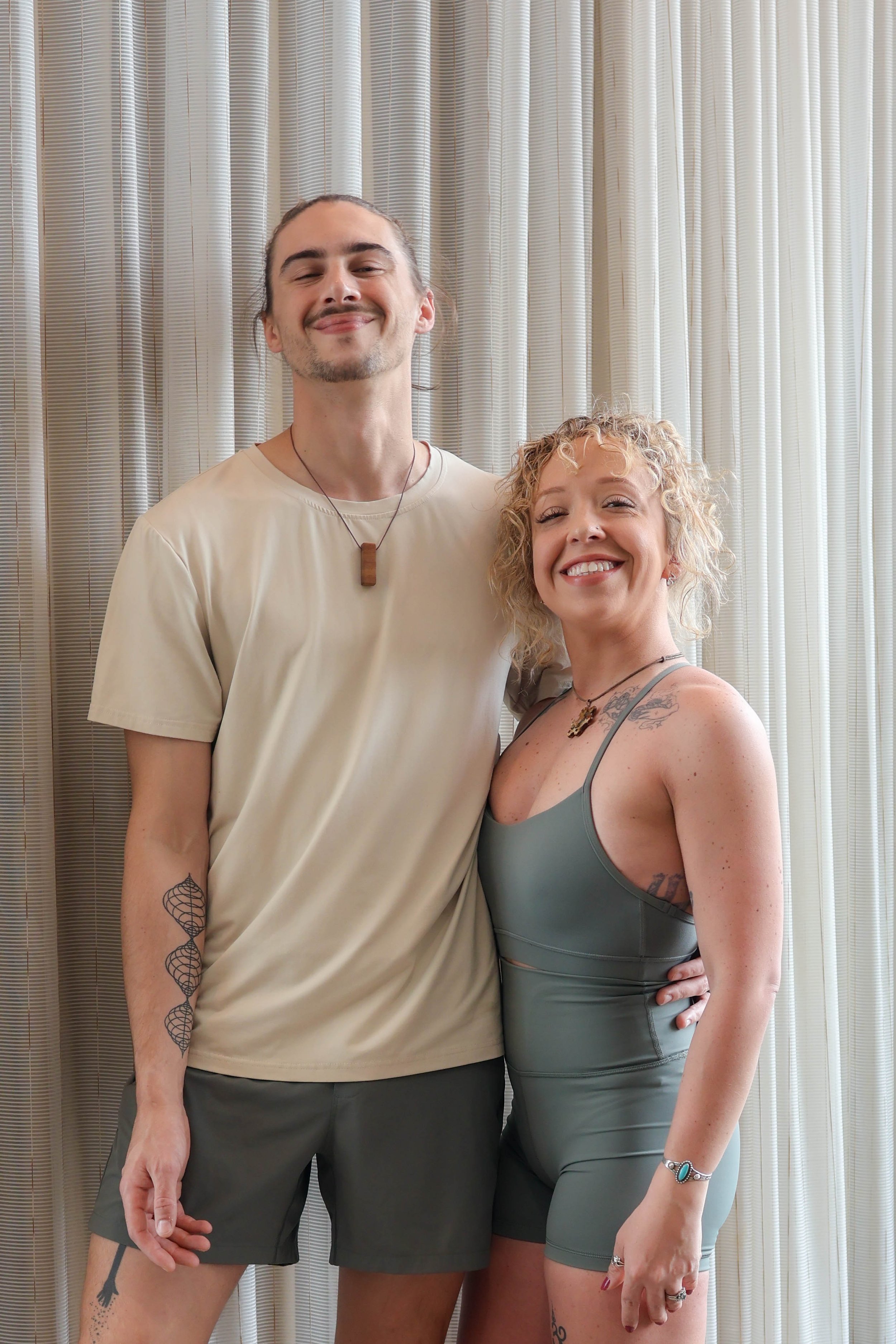 A smiling man with long hair pulled back and a mustache, wearing a beige t-shirt, dark shorts, and a wooden pendant necklace, standing next to a smiling woman with curly blonde hair, tattoos, wearing a gray tank top, and jewelry, with a curtain in the background.