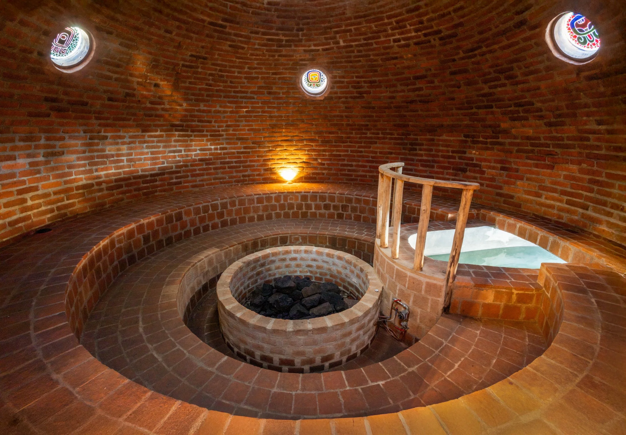 Interior of a red brick circular room with small round windows decorated with colorful tribal or indigenous patterns. There is a circular brick fire pit in the center filled with charcoal or rocks, and a curved brick seating area surrounding it. A wo