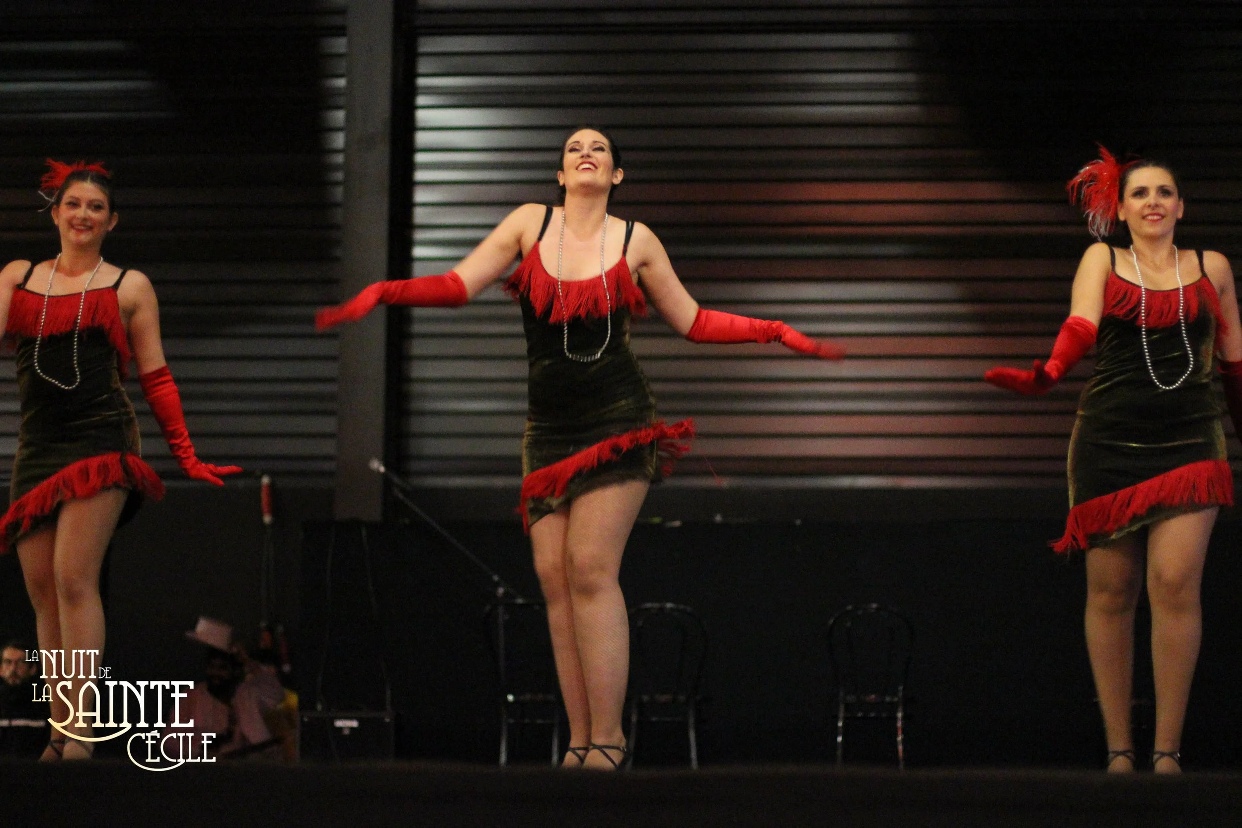 Trois femmes dans des costumes de cabaret rouge et noir, portant des gants rouges, se produisent dans un spectacle de danse au théâtre.