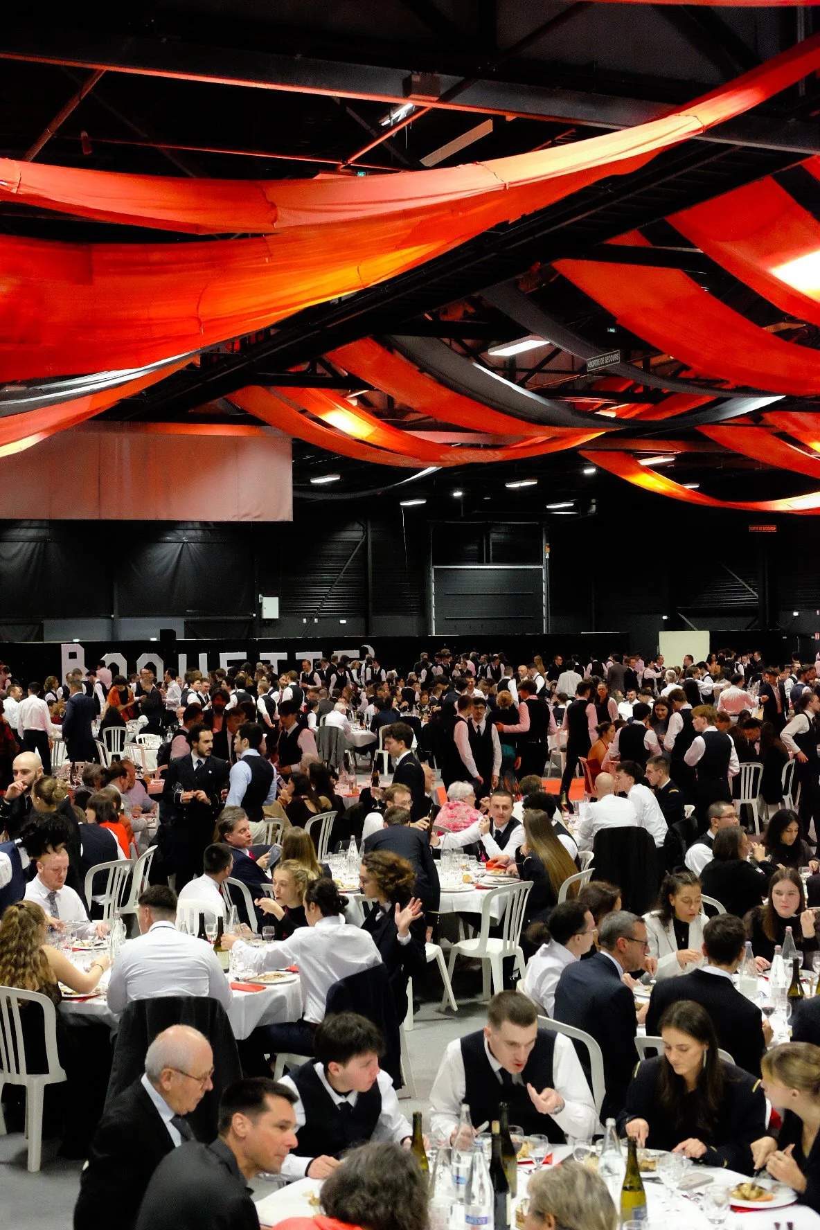 Une grande salle avec de nombreux convives à un repas, des serveurs en uniforme noir et blanc, des décorations en tissu orange suspendues au plafond.