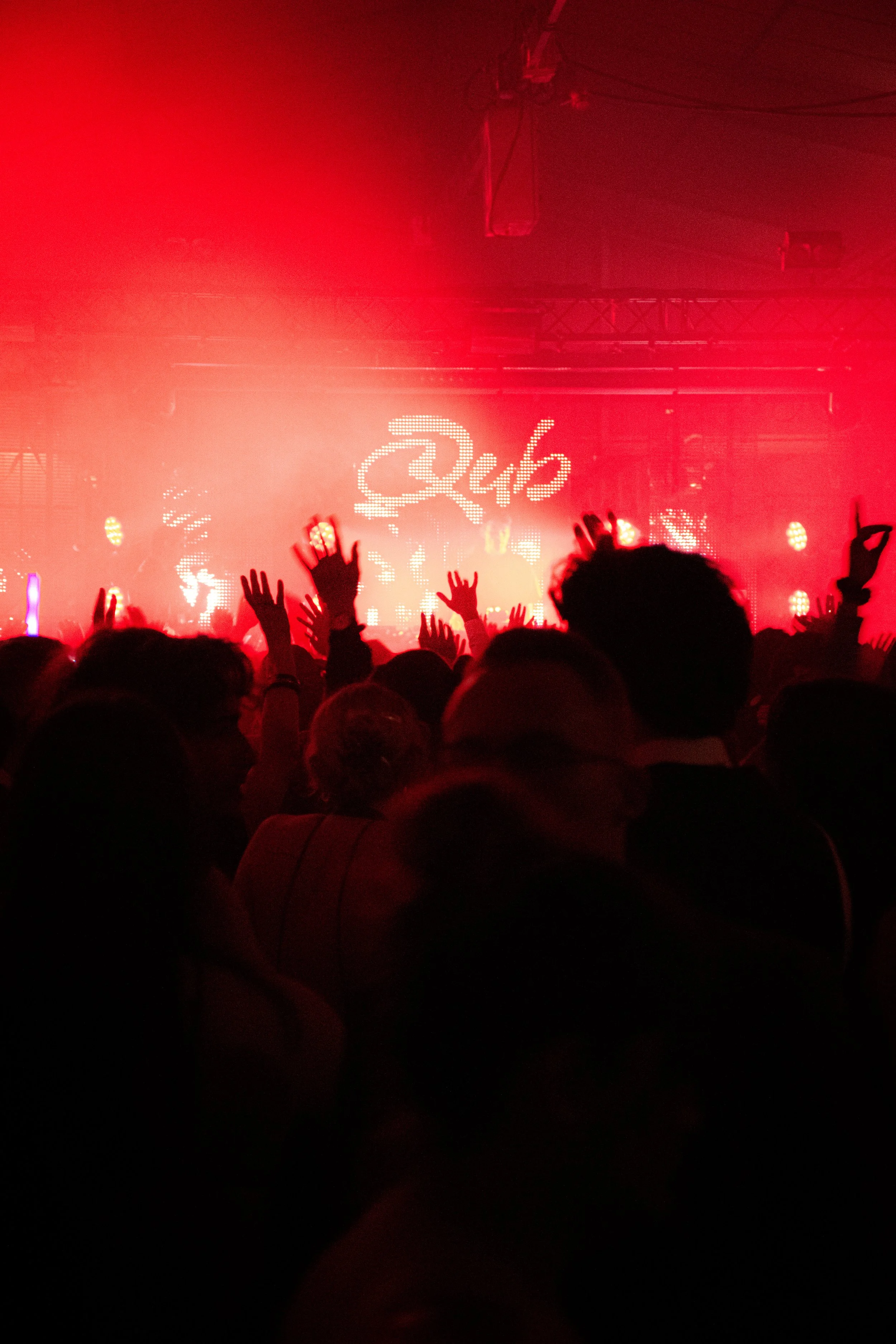 Une foule de personnes lors d'un concert ou d'un événement, avec des mains levées, sous un éclairage rouge et un écran LED en arrière-plan affichant le mot 'Qub'.