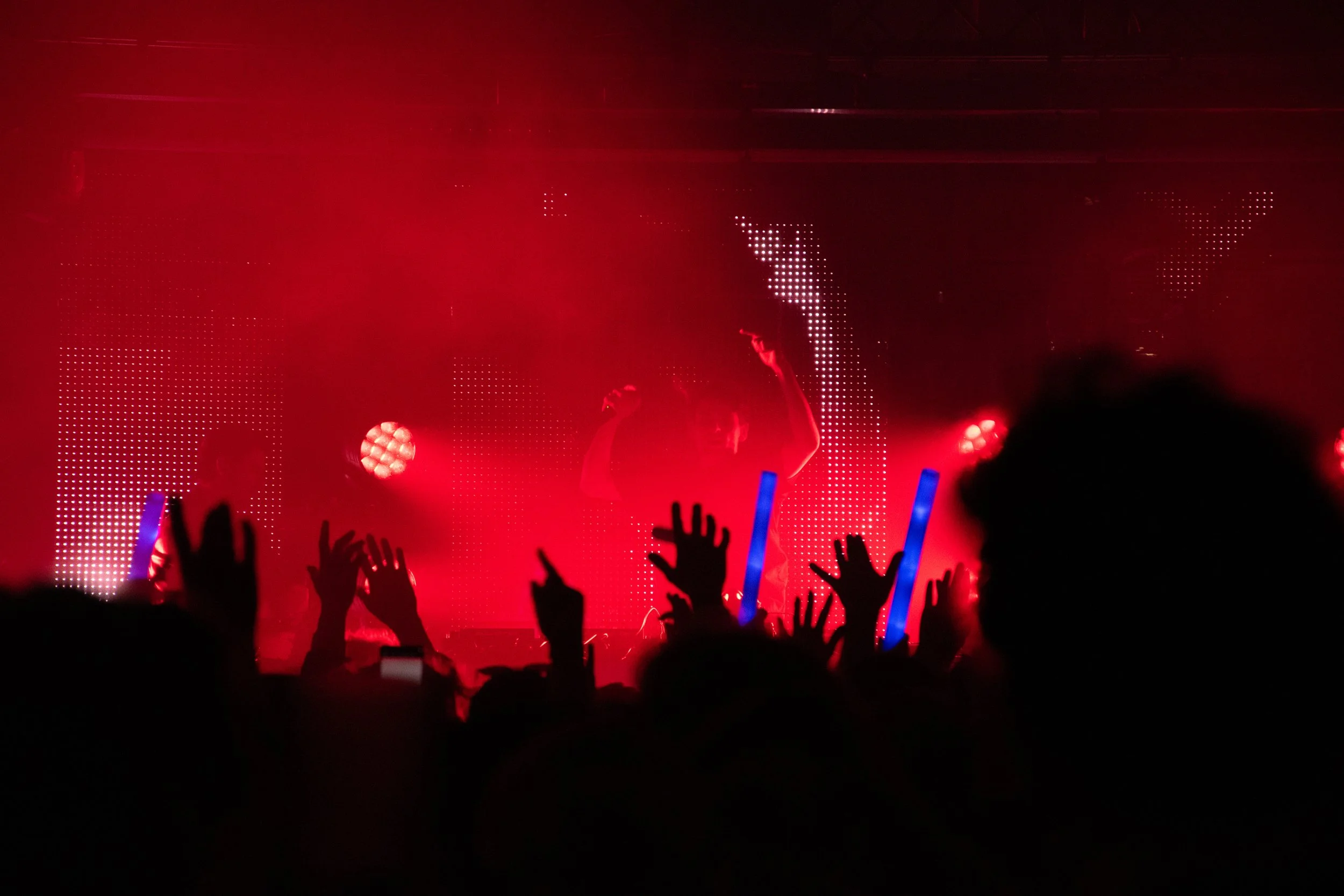 Un spectacle en direct avec des silhouettes de personnes dansant ou applaudissant, éclairé par des lumières rouges et des éléments lumineux bleus, avec un fond lumineux et des mains levées dans la foule.