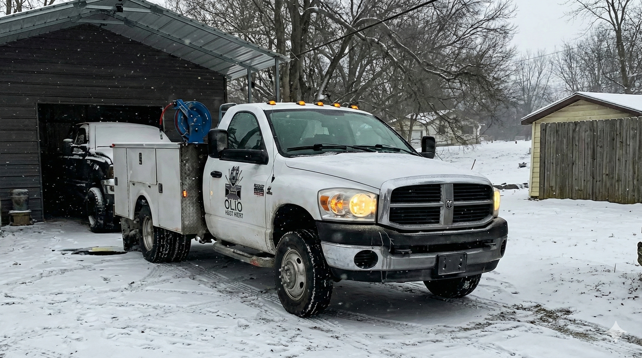 A white utility truck with a hose reel on the side is parked in a snow-covered driveway next to a black garage. The truck has a logo with wrenches and the words "Hollenbeck Fleet Services". The background shows leafless trees, snow on the ground.