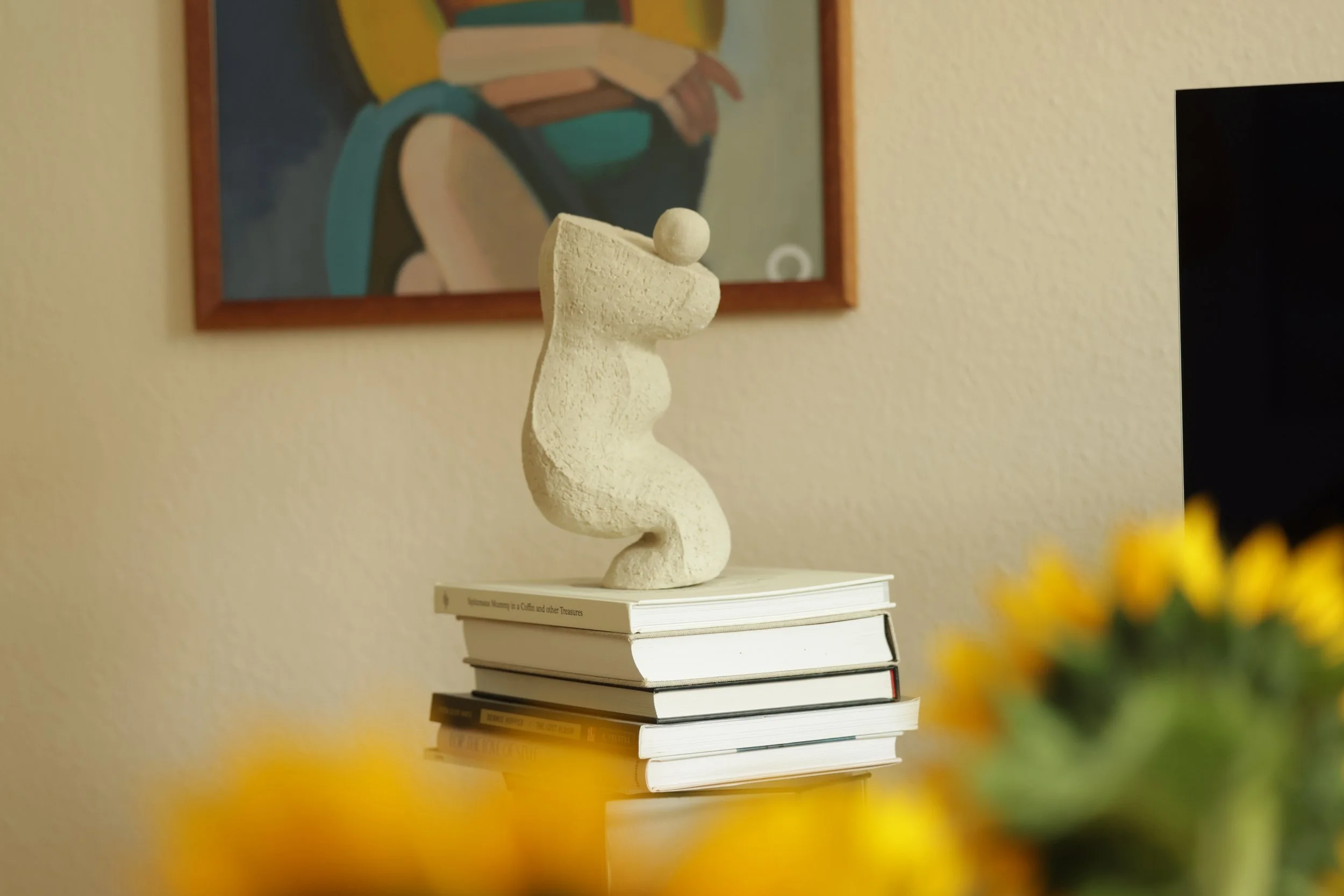 Eine Skulptur einer abstrakten Figur, die auf einem Stapel Bücher auf einem weißen Regal steht. Im Hintergrund ein gemaltes Bild mit Frauen übrigens in Gelb und Blau. Die Szenerie ist in warmen Tönen gehalten.