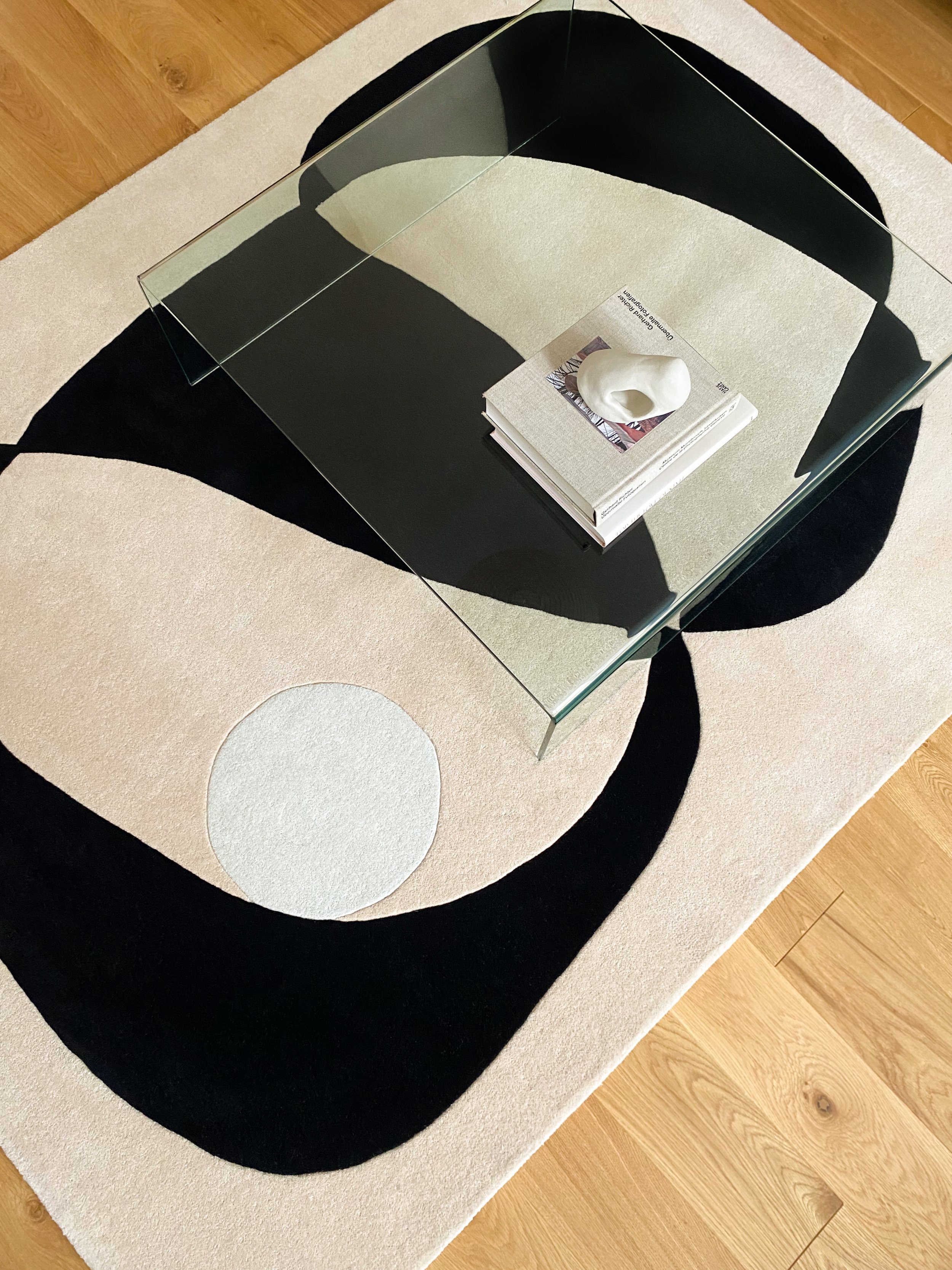 Ein Tisch mit einem Glasdeckel, darunter befindet sich ein aufgeschlagenes Buch und eine weiße Skulptur auf einem kleinen Magazin, der Tisch steht auf einem schwarzen und weißen ovalen Teppich, der auf Holzboden liegt.