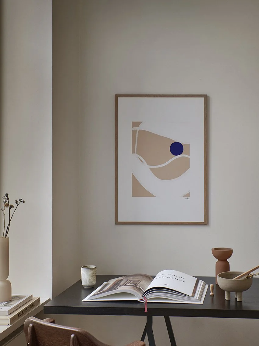 Ein Schreibtisch mit offenen Büchern, Kerzen und Keramikvasen vor einer abstrakten Kunstzeichnung an der Wand.