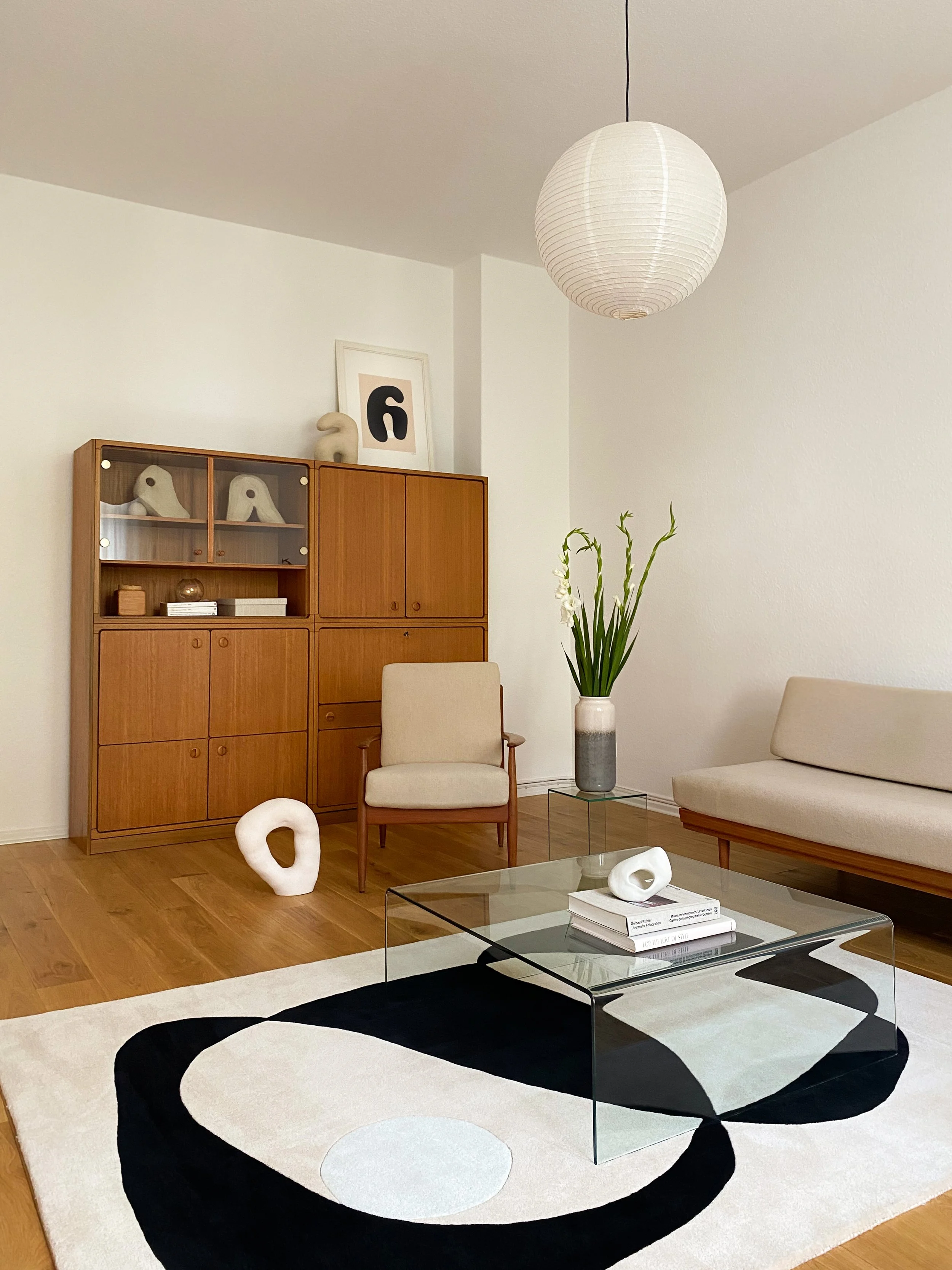 Ein modernes Wohnzimmer mit einem weißen Sofastuhl, einem Glastisch, einem großen schwarzen und weißen Teppich mit geometrischem Muster, einem Bücherregal aus Holz mit Dekorationsgegenständen und einem Pflanzenstrauch. An der Wand hängen Bilder und eine Papierlampe an der Decke.
