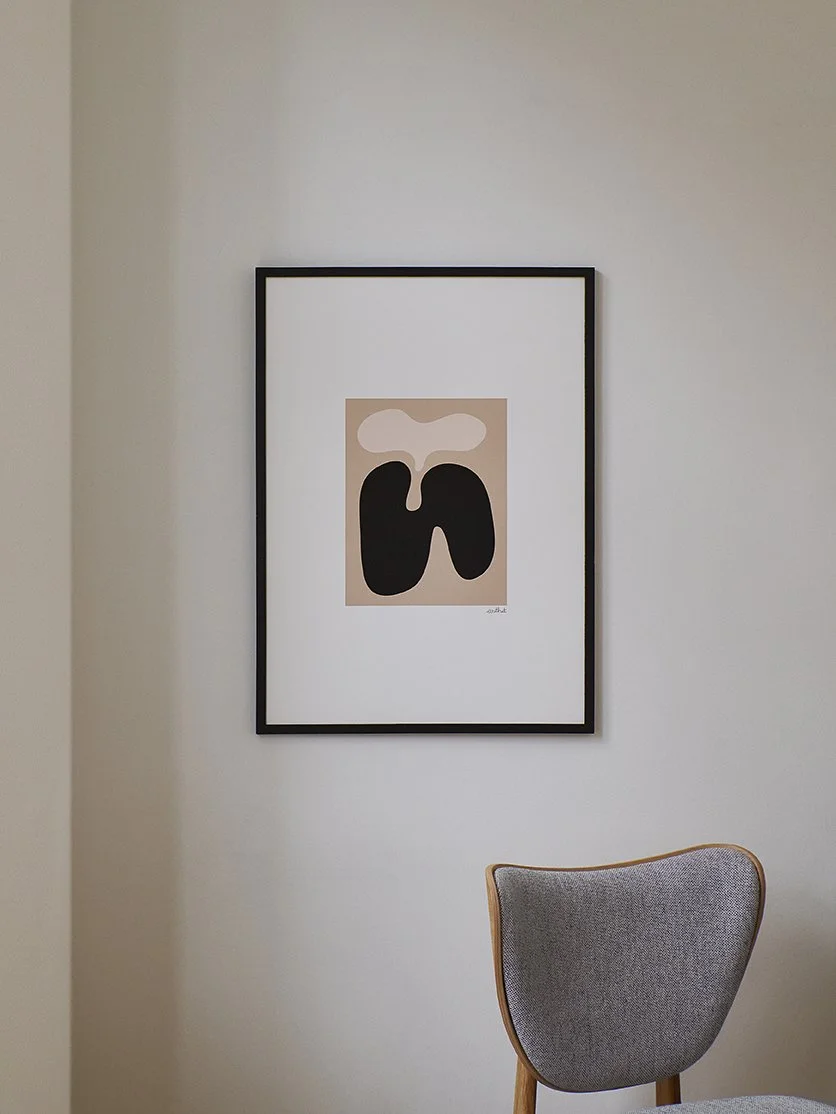 Abstraktes Kunstwerk mit schwarzen und weißen Formen, im Rahmen an einer weißen Wand, daneben ein grauer Stuhl.