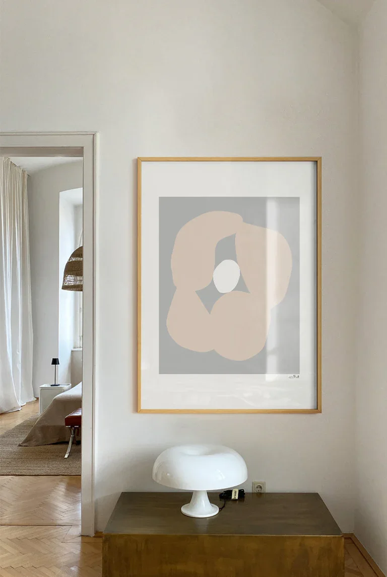 Ein ruhiger Raum mit weißer Wand, auf der ein abstraktes Kunstwerk mit neutralen Tönen hängt. Darunter steht ein Holztablett mit einer weißen Lampe.