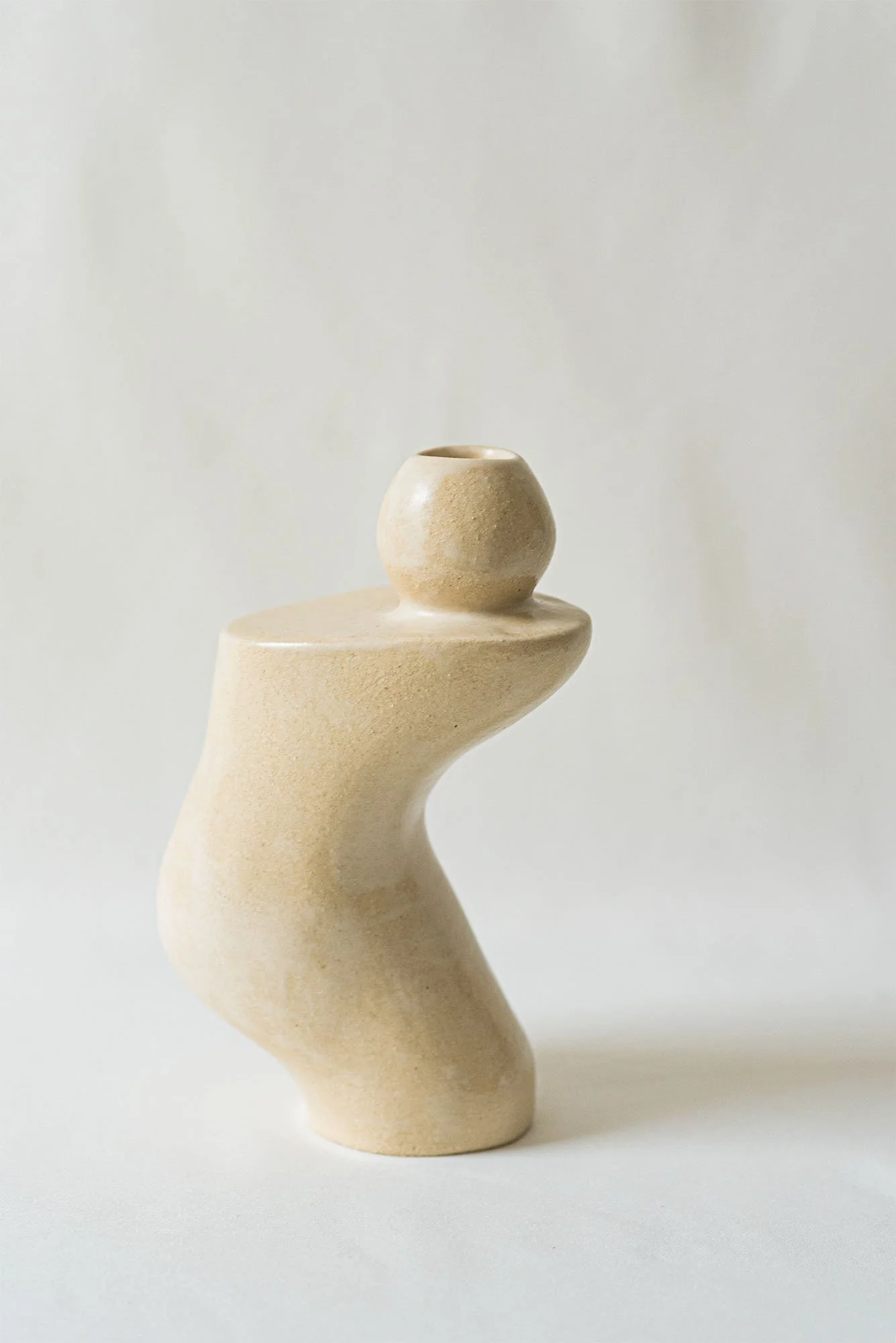 Skulptur aus hellem, glattem Material, die die Form eines abstrakten, stilisierten Vogels oder einer Figur mitentwickelten, organischen Form hat, steht auf einer weißen Oberfläche gegen einen neutralen Hintergrund.