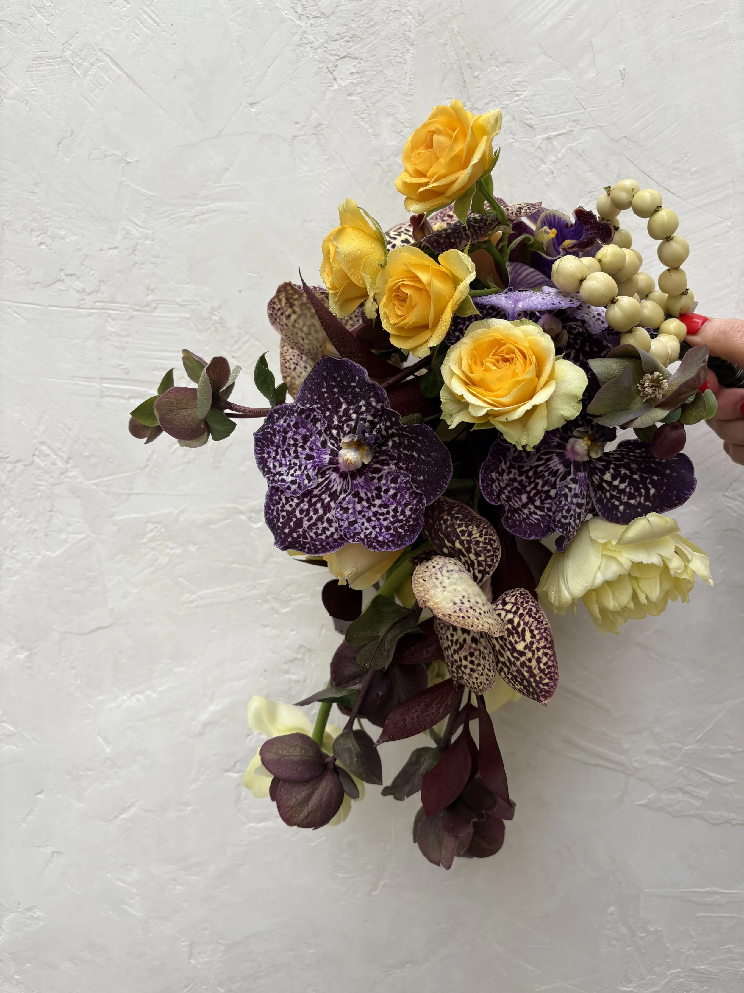 A bouquet of yellow roses, purple orchids, white berries, and green leaves held by a hand with red-painted nails against a white textured wall.