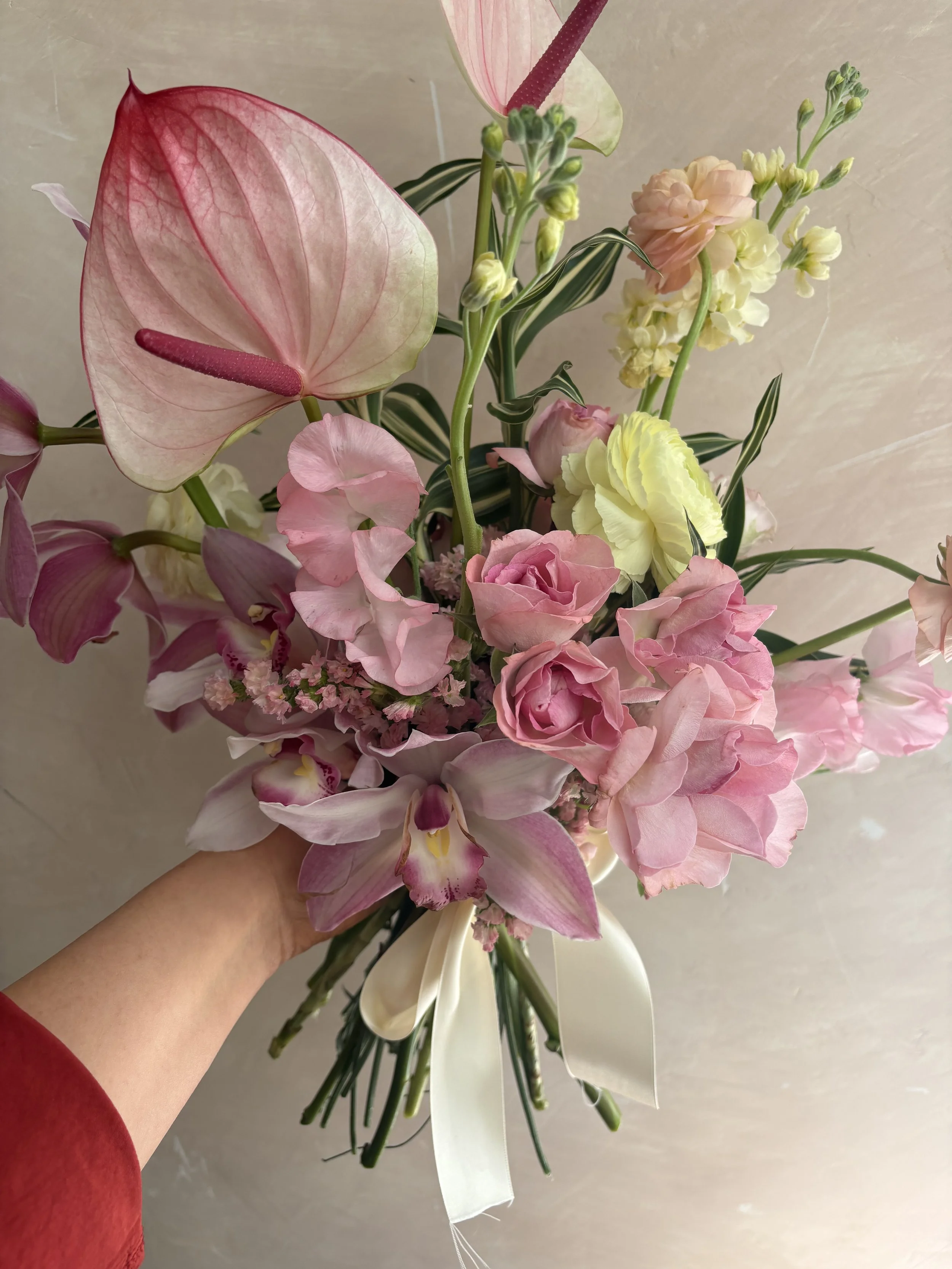 A hand holding a bouquet of various pink and white flowers including anthuriums, roses, and other blooms, tied with a white ribbon.
