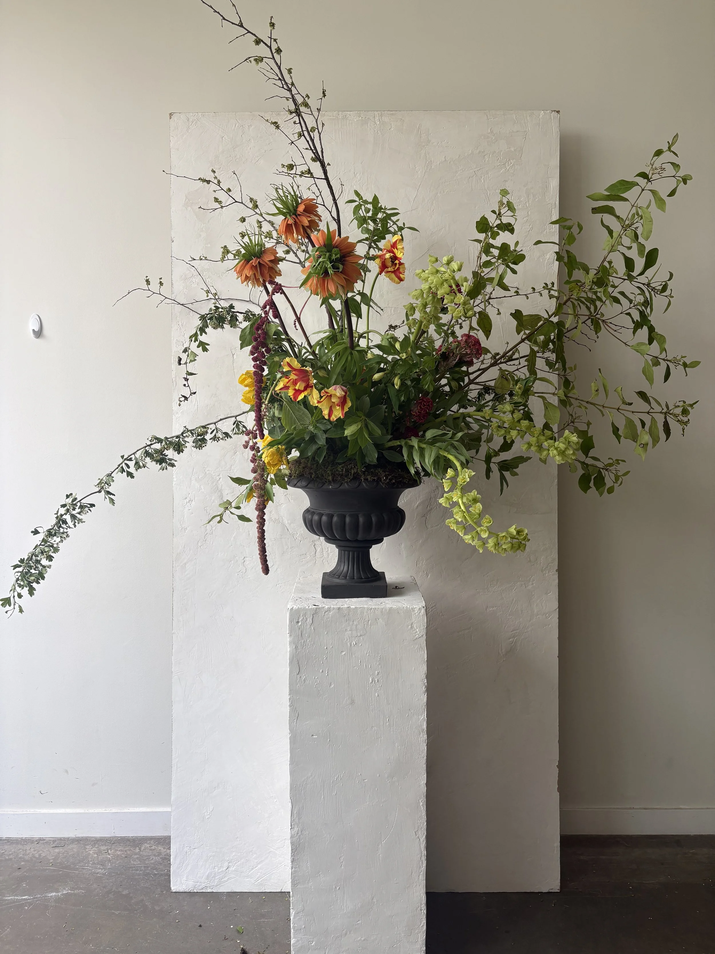 A black pedestal vase with an arrangement of mixed flowers and branches, set against a white textured wall and a white rectangular pedestal.