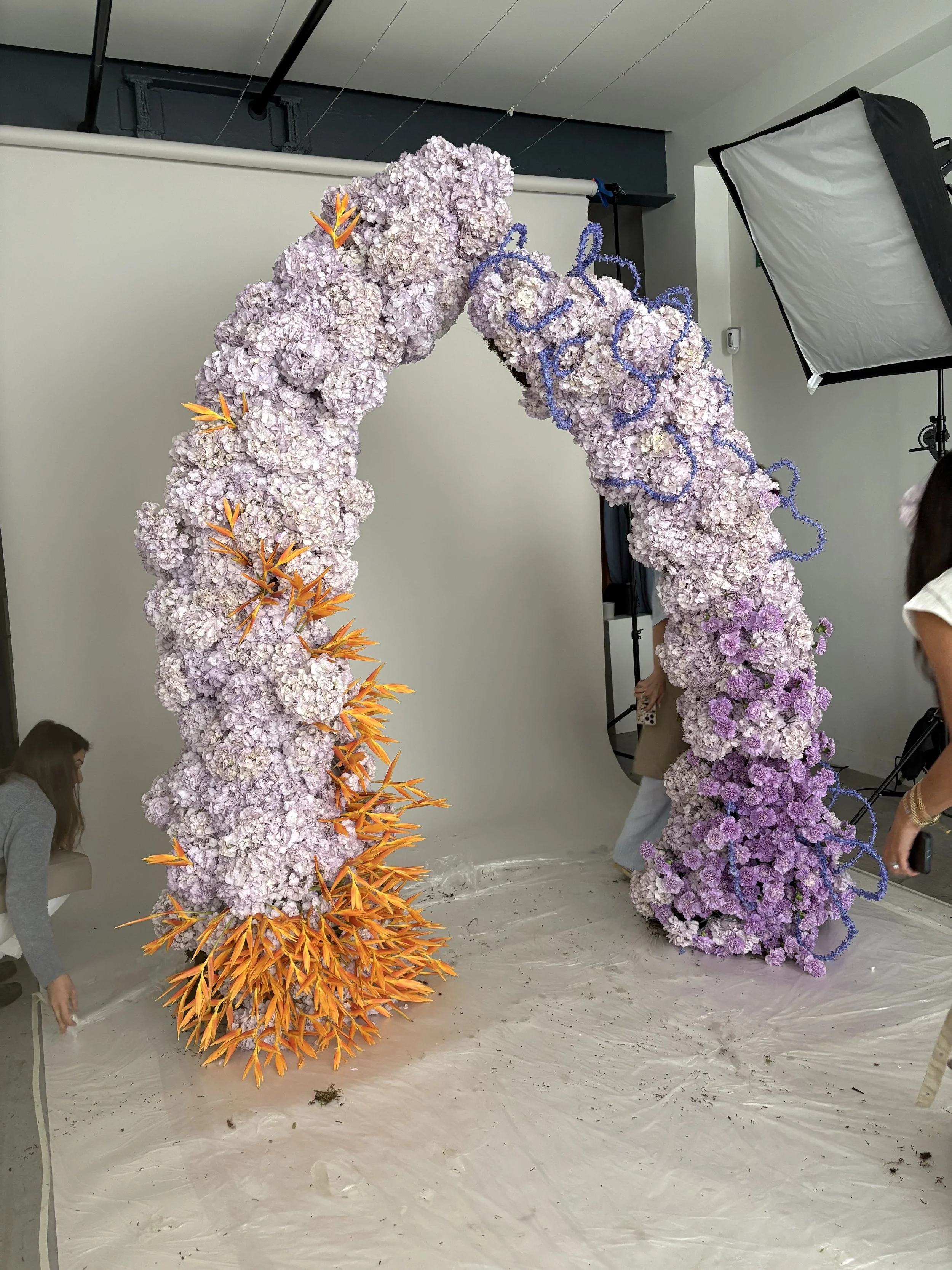 Large floral arch made of purple and light pink flowers with orange and blue floral accents, set up in a photography studio with people adjusting it.