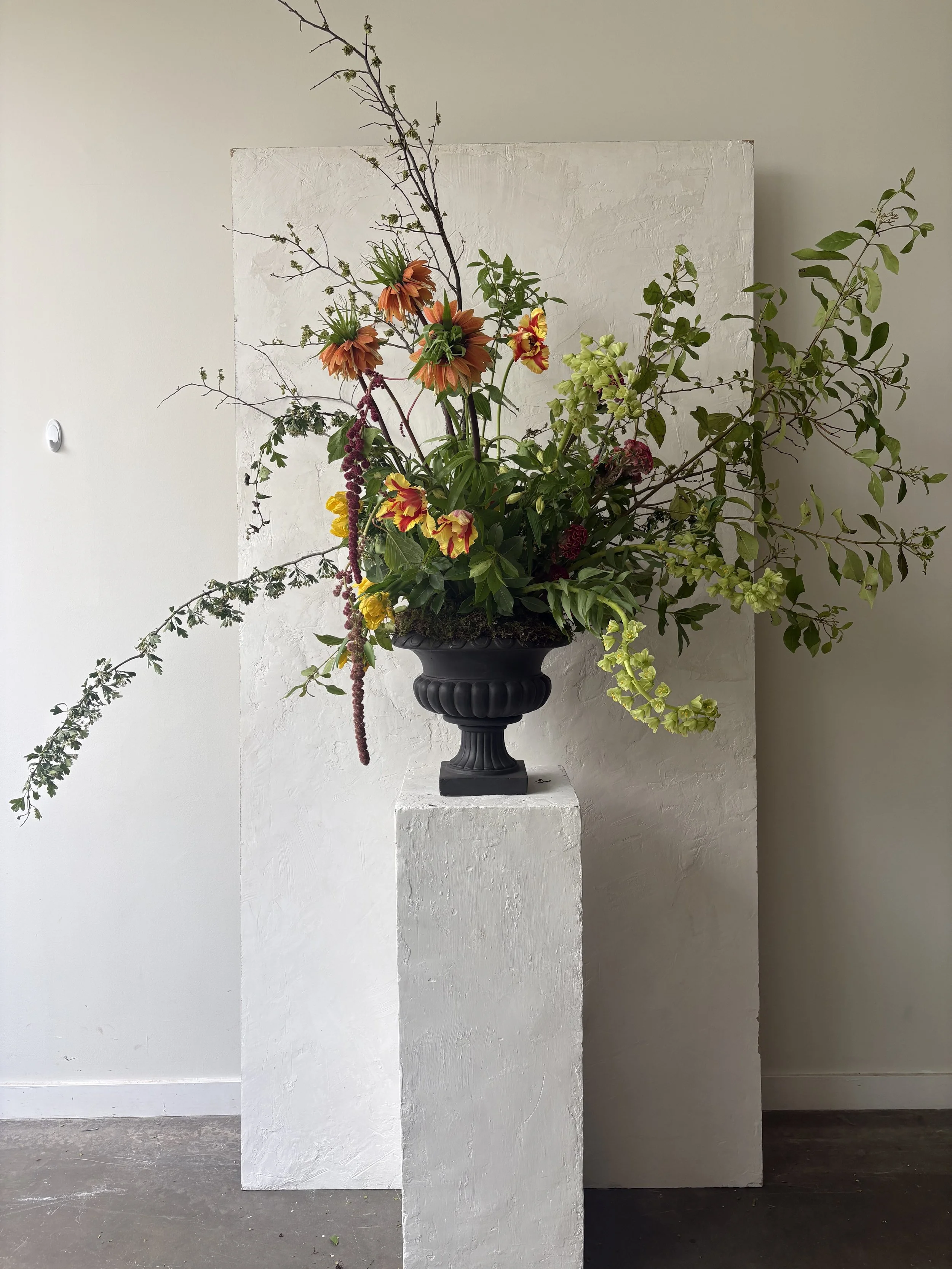 A large floral arrangement in a black vase, placed on a white pedestal against a textured white wall.