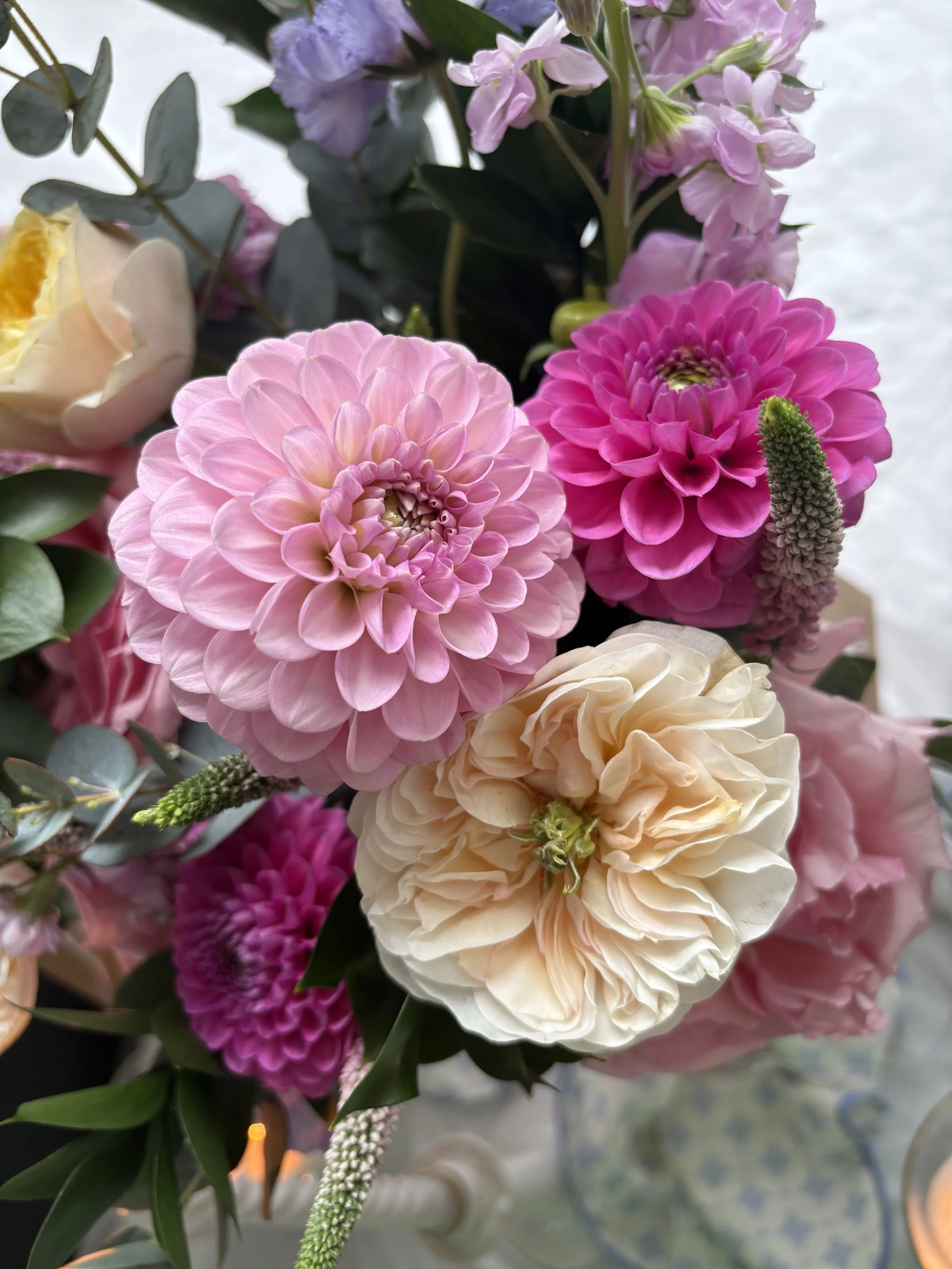 Close-up of a vibrant bouquet featuring light pink and deep pink dahlias, white roses, hydrangeas, and green foliage, with a blurred background.