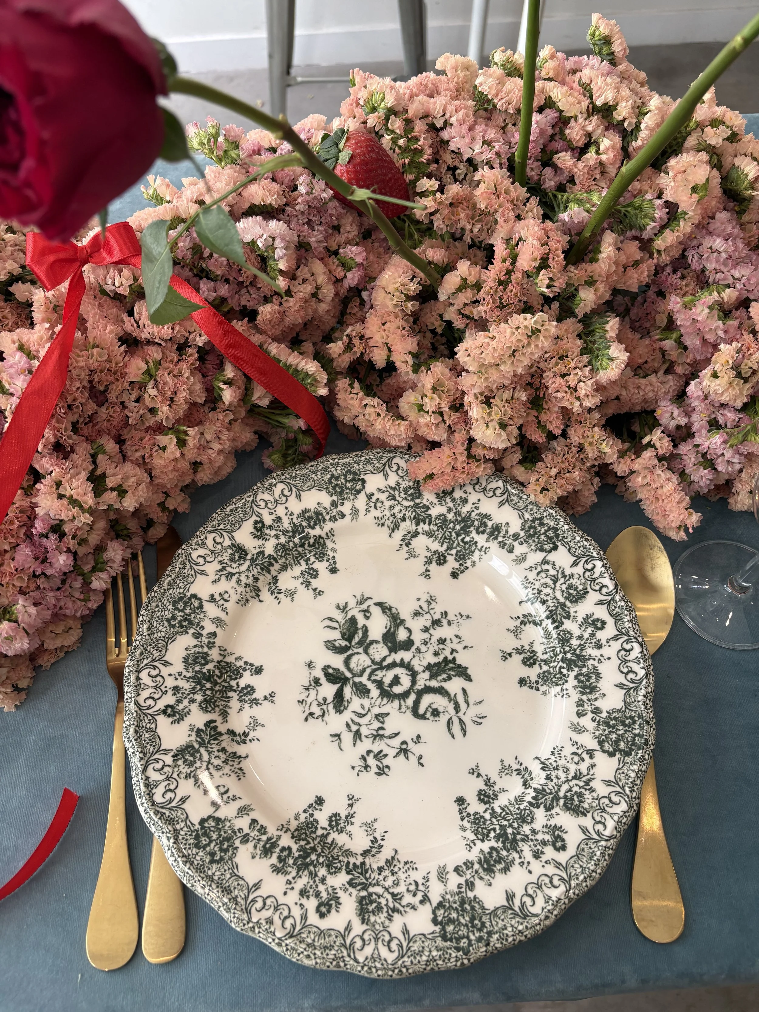 A table setting with a decorative plate that has a black floral pattern, gold cutlery, and a bouquet of pink flowers, including a partially visible red rose, strawberry, and a single tulip on a dark blue tablecloth.