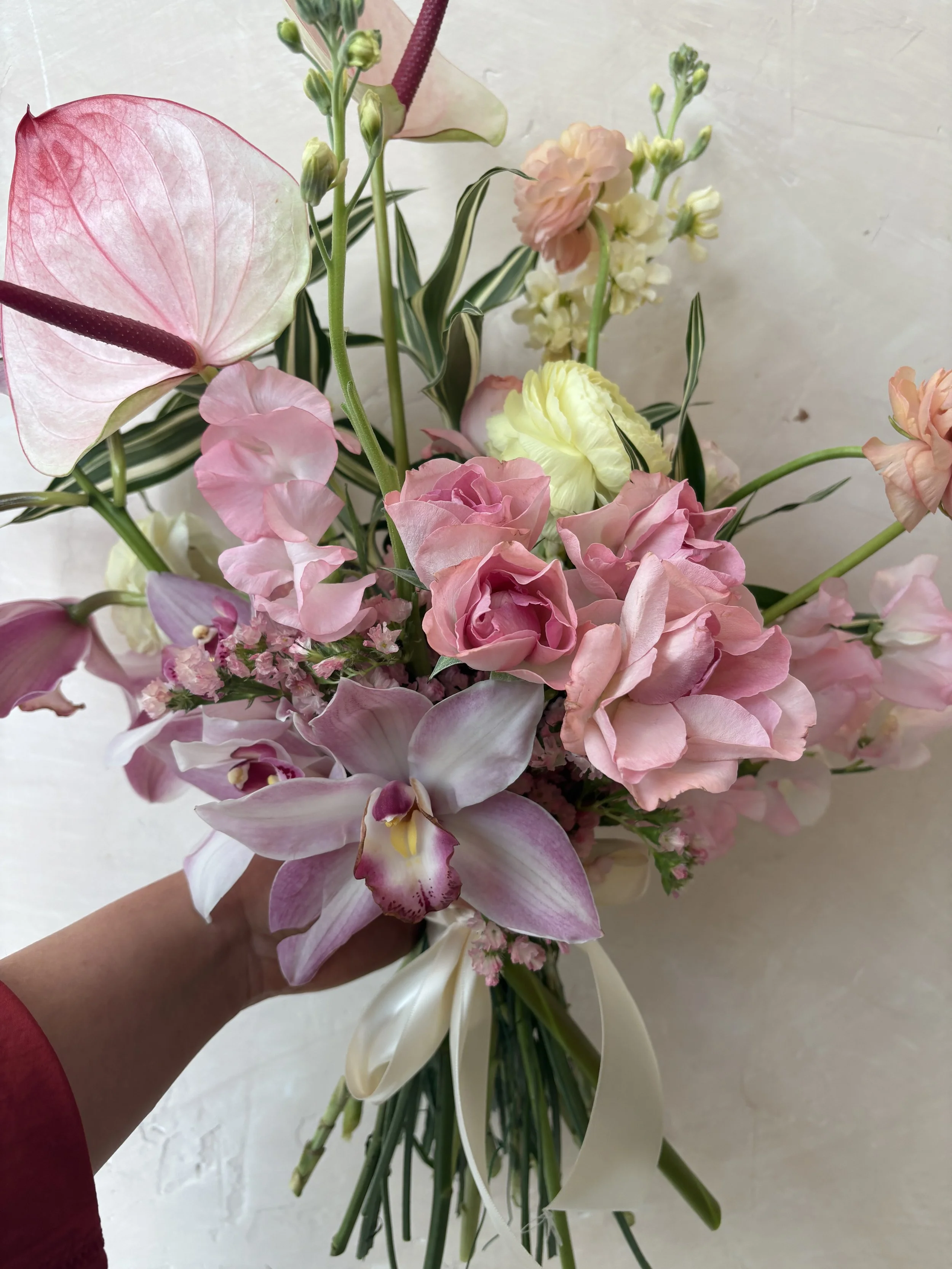 A hand holding a bouquet of pink, white, and peach flowers with green stems and leaves on a white surface.