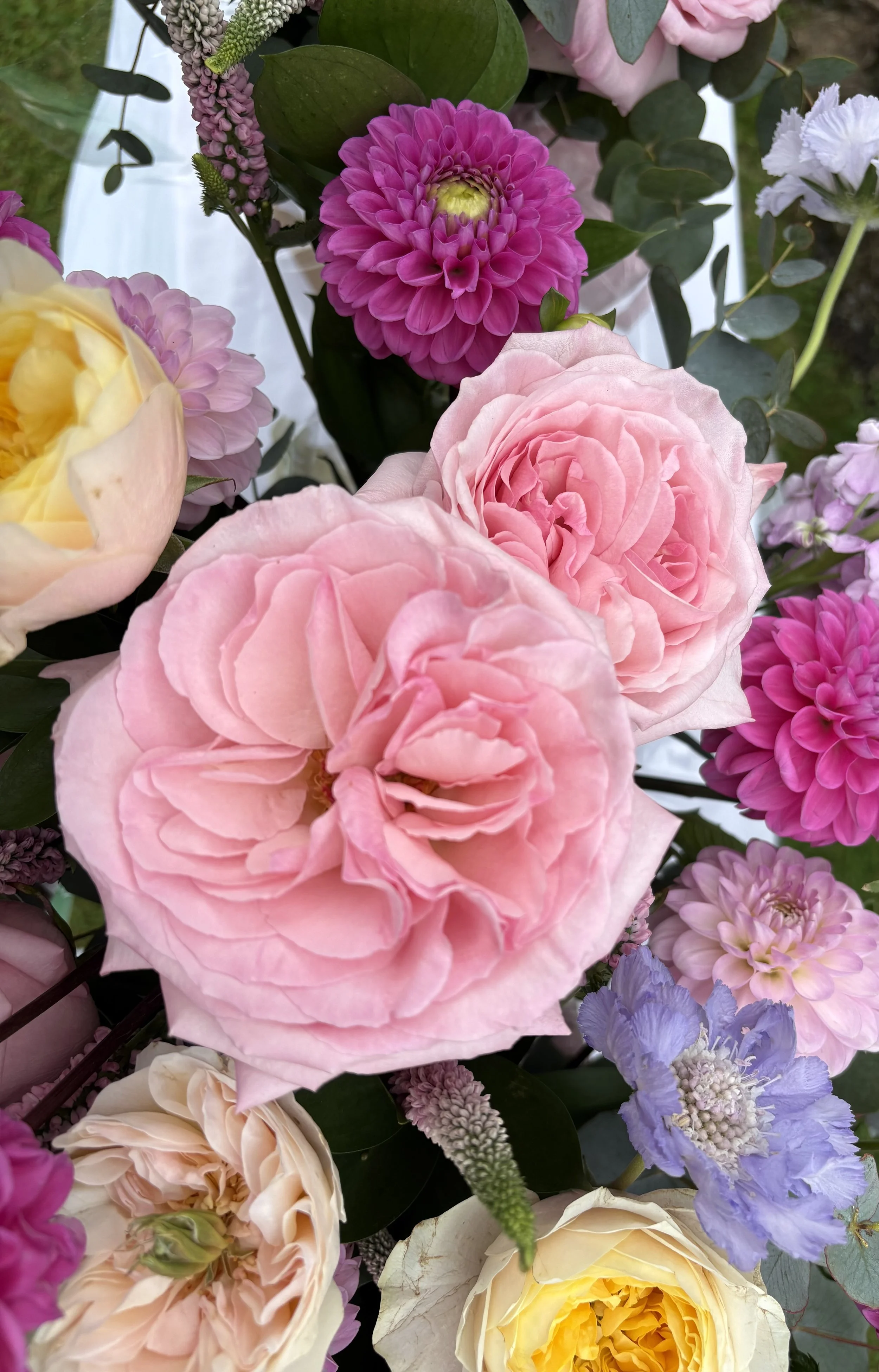 A bouquet of various colorful flowers including pink dahlias, yellow roses, purple lisianthus, and other blooms with green leaves.