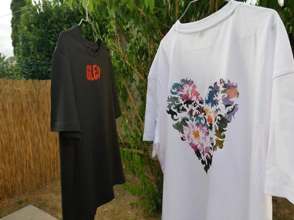 Deux T-shirts oversize en coton lourd, un noir et un blanc, suspendus à une corde à linge. On voit le logo "GLEAD" orange face avant et le design cœur avec inspiration tableau Monet face arrière.