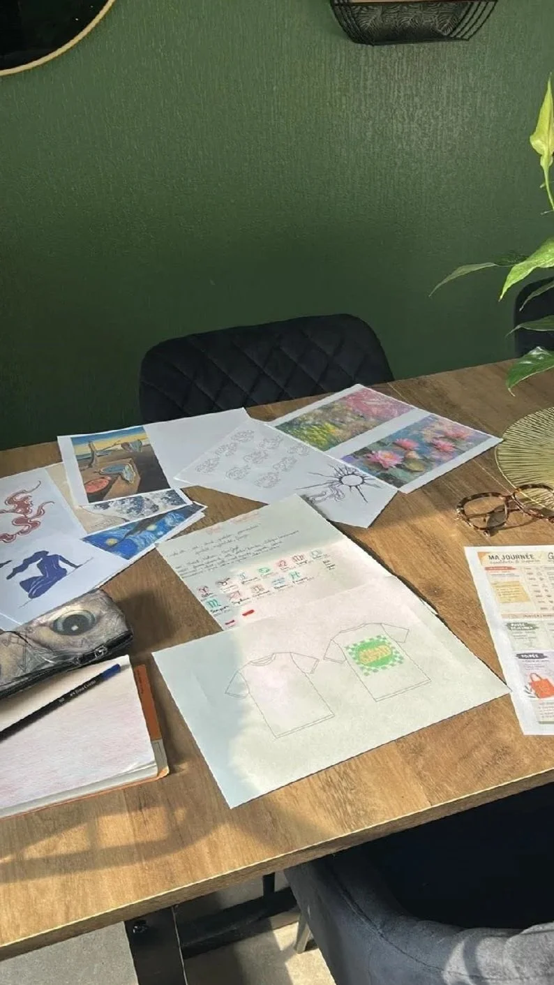 Table de travail avec des feuilles de papier montrant des croquis et des inspirations, des dessins, un carnet, un stylo, une paire de lunettes, un magazine, et quelques plantes. Marque de streetwear créée à Albi