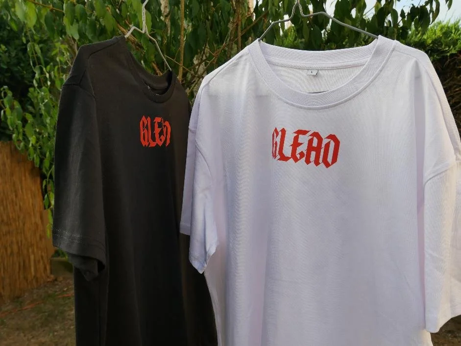 Deux vêtement streetwear artistique coupe loose en coton lourd, un noir et un blanc, suspendus à une corde à linge. On voit le logo "GLEAD" orange, placé face avant, pour chaque t-shirt.