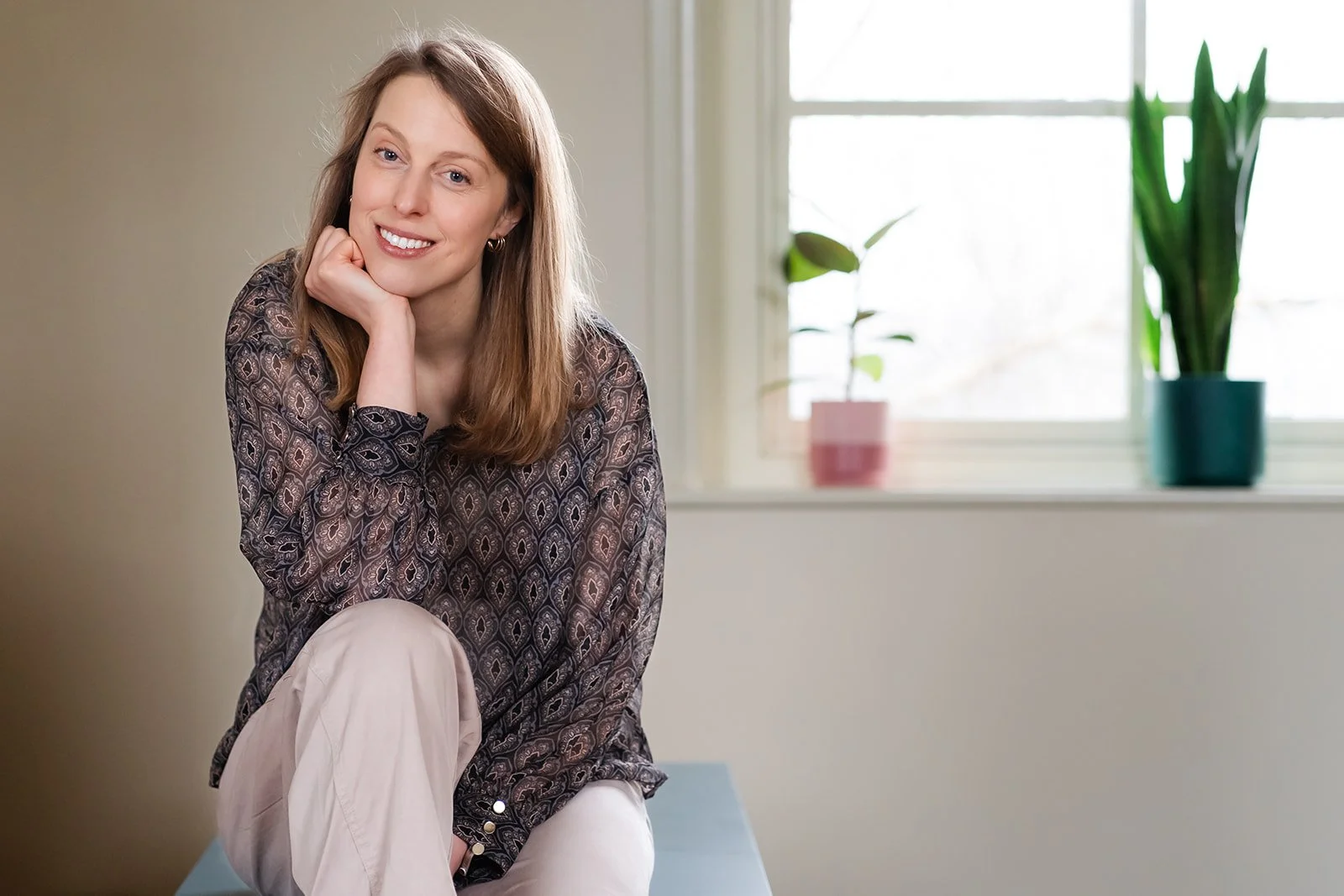 A woman with brown hair, wearing a patterned blouse and light-colored pants, sitting indoors near a window with potted plants on the windowsill.