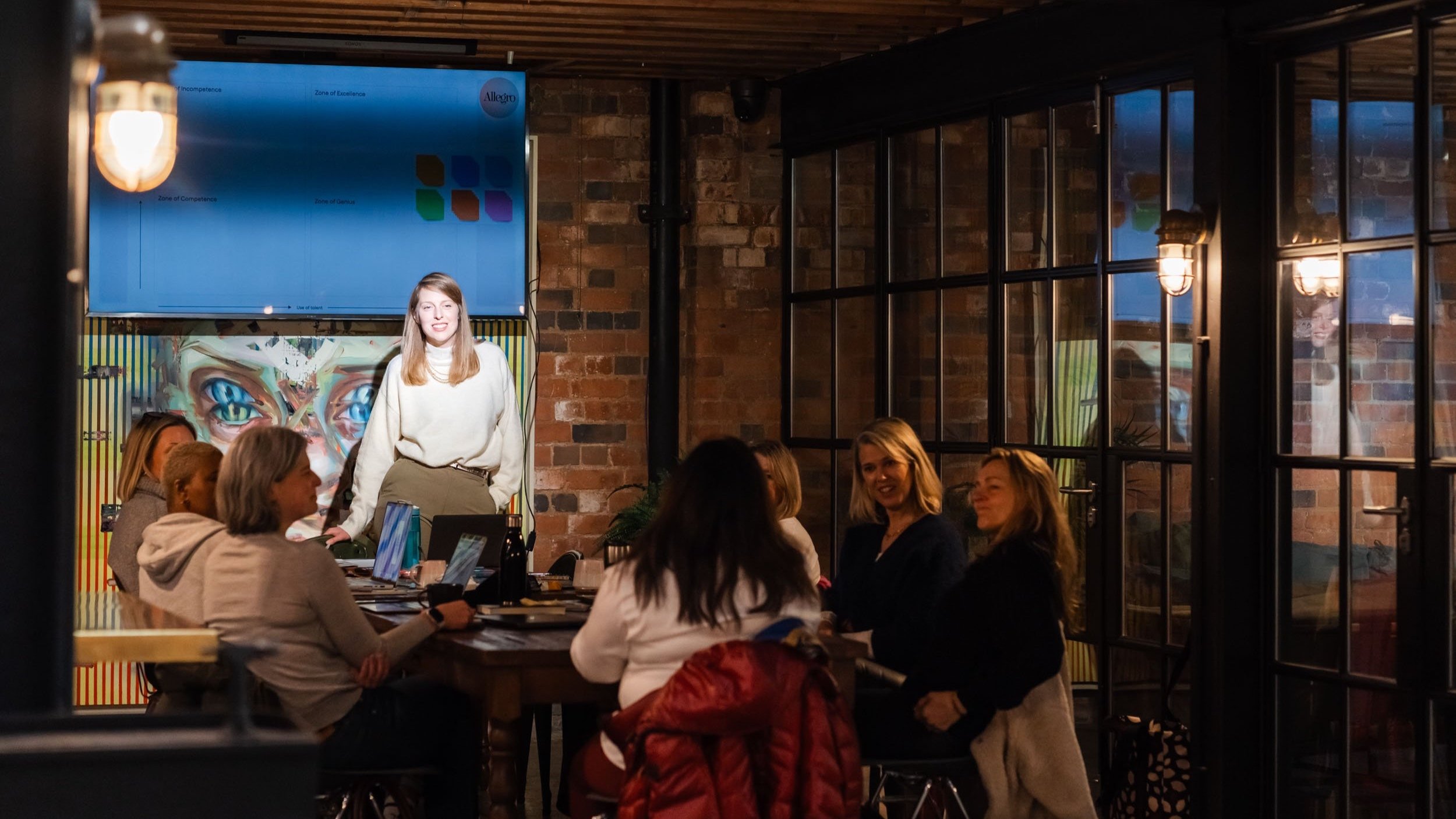 A group of people socializing and working on laptops in a cozy, brick-walled restaurant or cafe with large glass windows and warm lighting. A woman stands near a presentation screen with the Allegro logo, smiling.