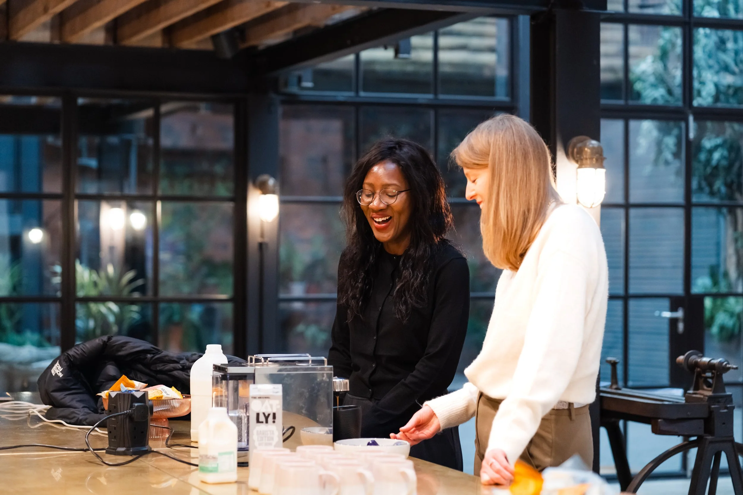 Two women are standing at a table inside a modern building with large windows and wooden ceiling beams, smiling and looking down at items on the table.