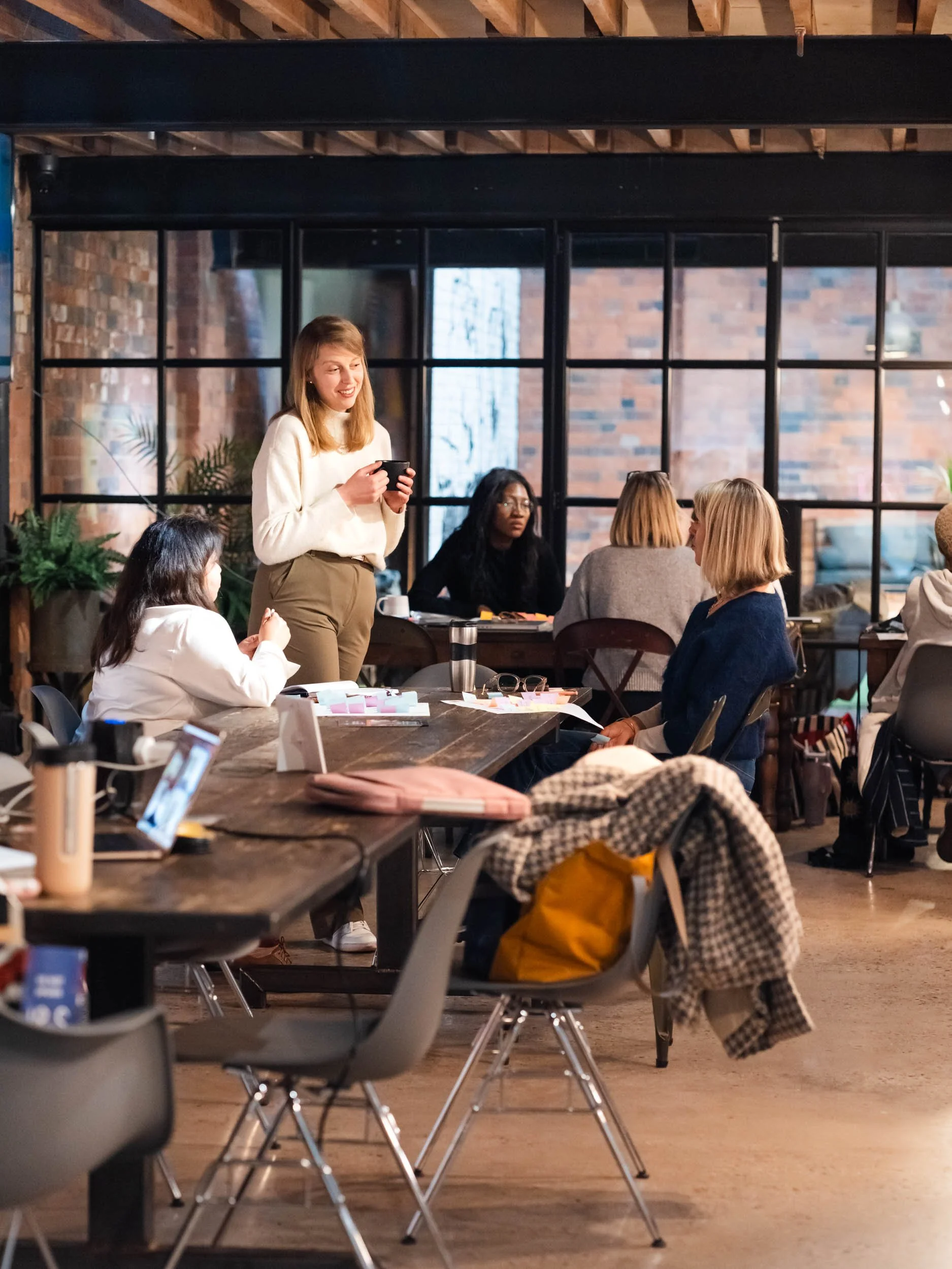 A group of diverse women's conference or workshop in a loft-style space with large windows, sitting at a long wooden table, engaging in discussion, with one woman standing and smiling while holding a mug.