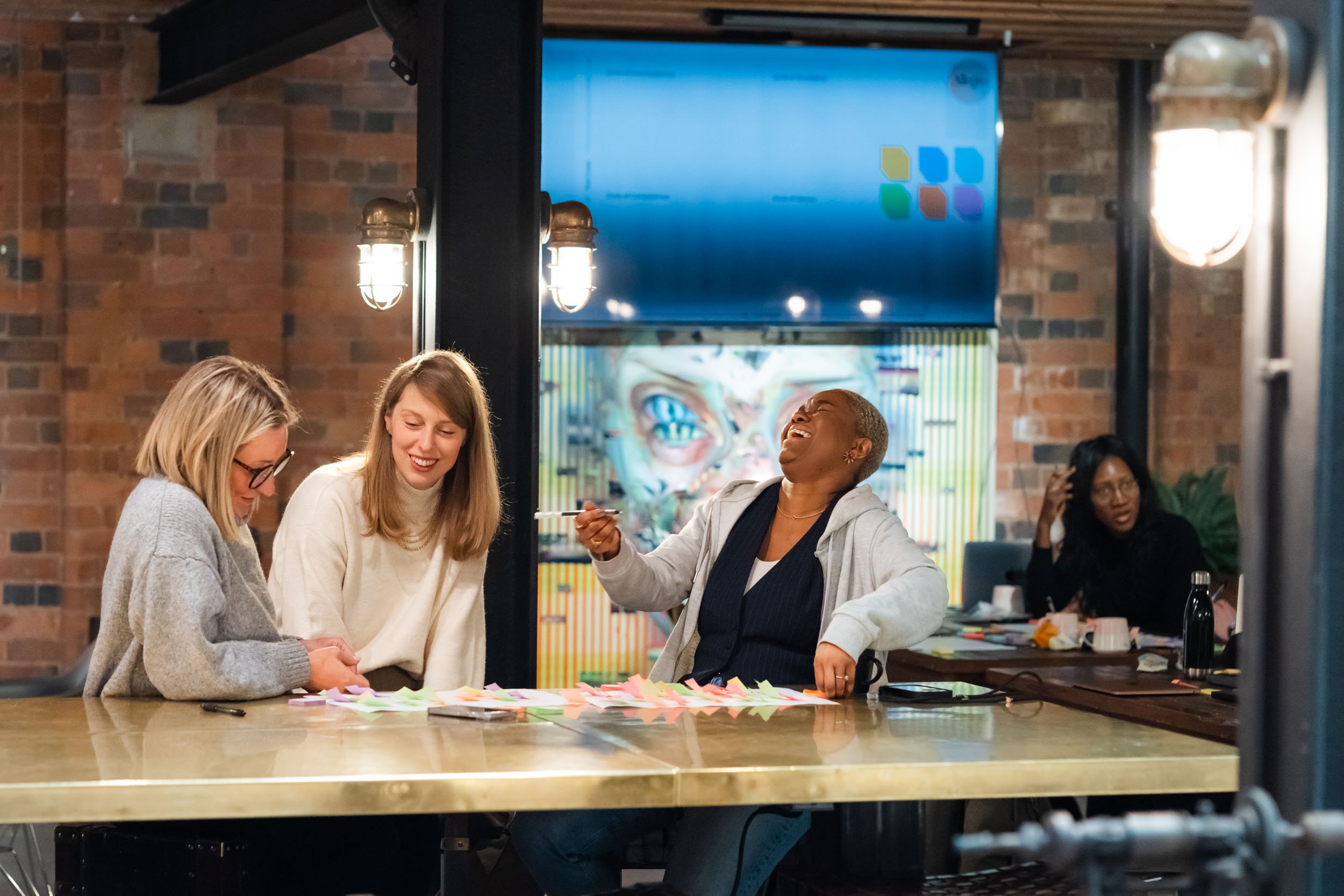 Group of women smiling and engaging in a discussion around a table with colorful paper notes in a modern office or coworking space.