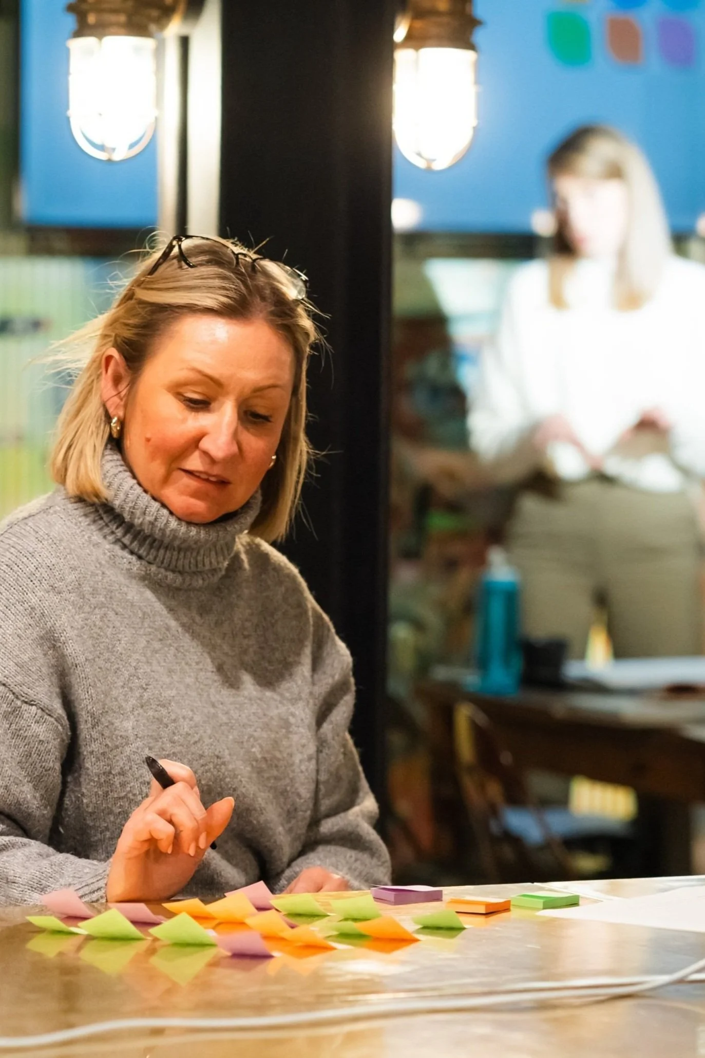 A woman with blonde hair wearing a gray turtleneck sweater is sitting at a table covered with colorful sticky notes. She is holding a black pen. In the background, another woman with light brown hair and glasses, dressed in white, is blurred and appears to be working. The setting seems to be a cozy indoor space with warm lighting.