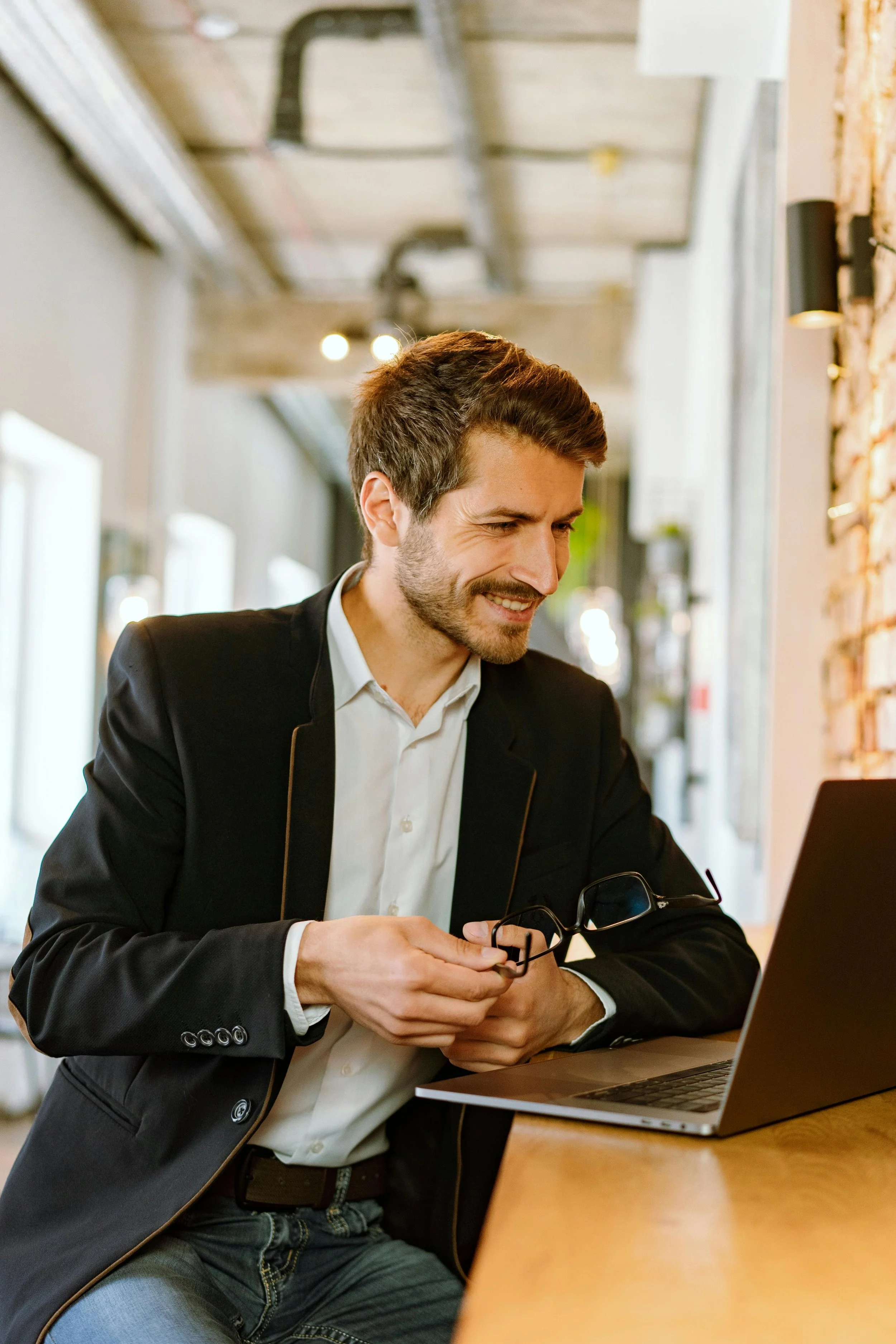 A man in a business suit smiling at a laptop, holding glasses, in a modern cafe with brick wall and large windows.