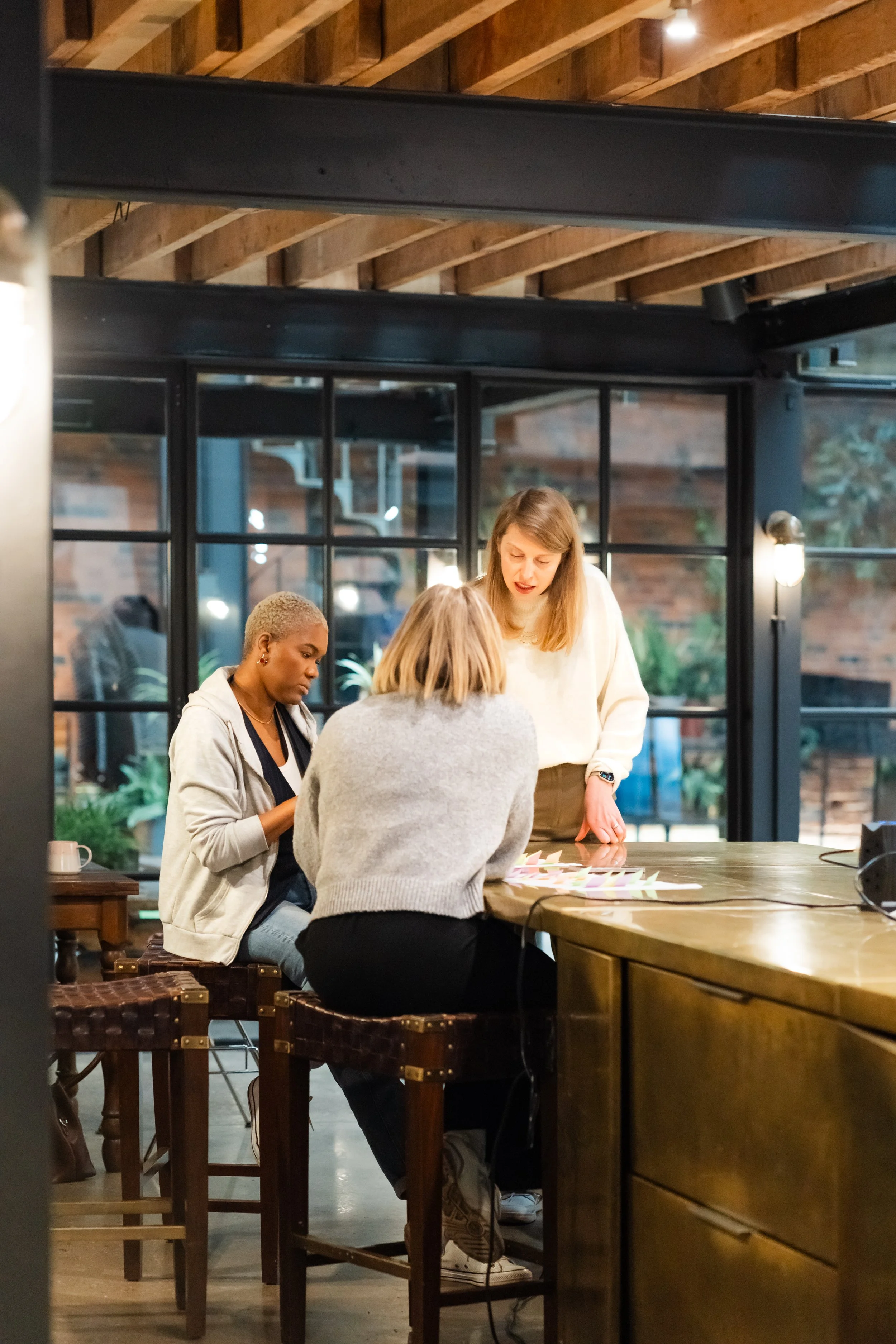 Four women sitting at a wooden table inside a modern restaurant with a brick wall visible outside through large glass windows, engaged in a discussion or activity.