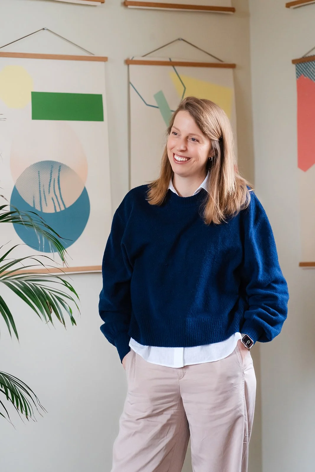 A woman with light brown hair, wearing a navy blue sweater, white shirt, and beige pants, standing inside a room with abstract colorful wall art and the plant on the left side.