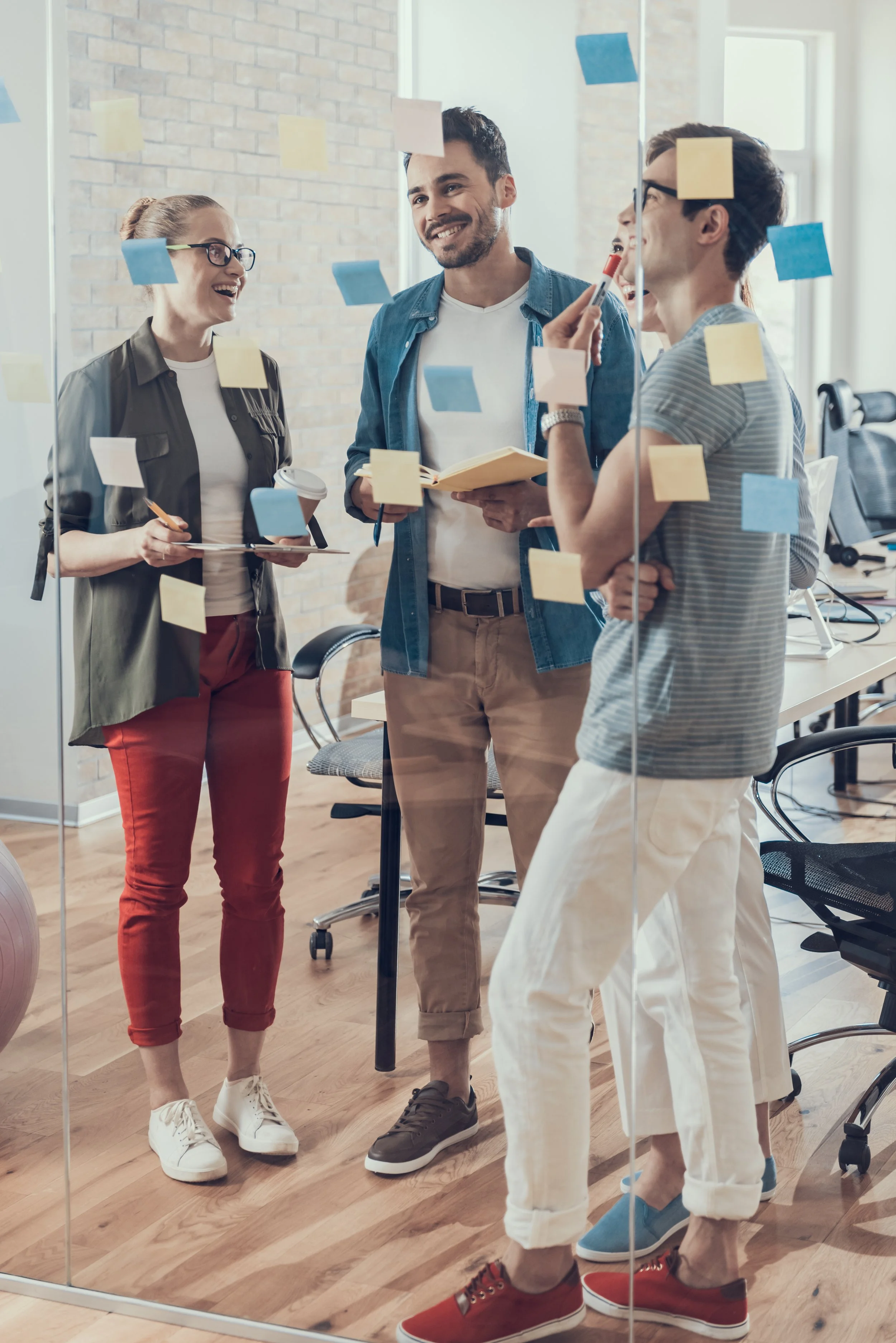 A diverse group of young professionals talking and collaborating in an office, seen through a glass wall with sticky notes.