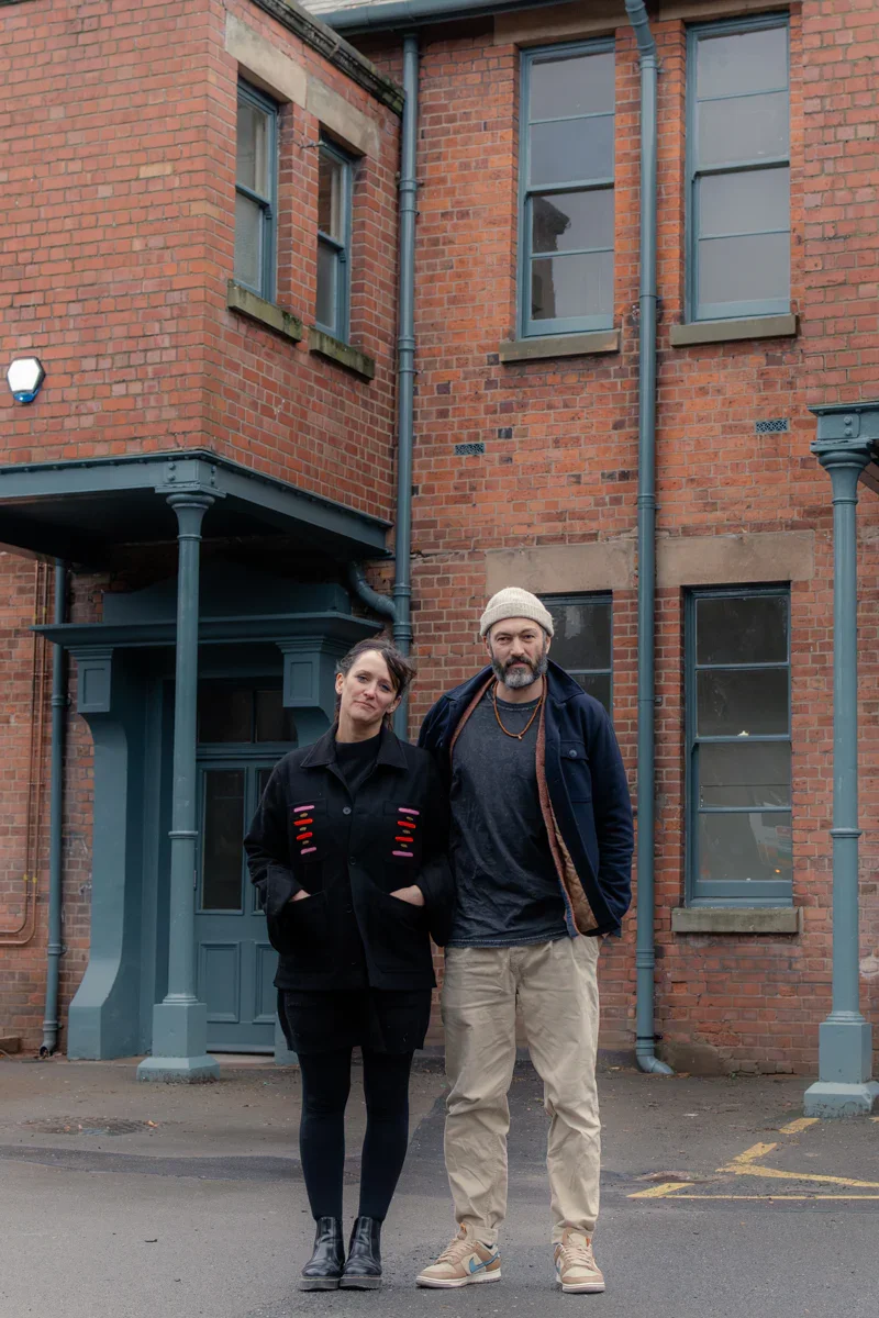 A woman and a man stand outside in front of a brick building with multiple windows and gray drainpipes.